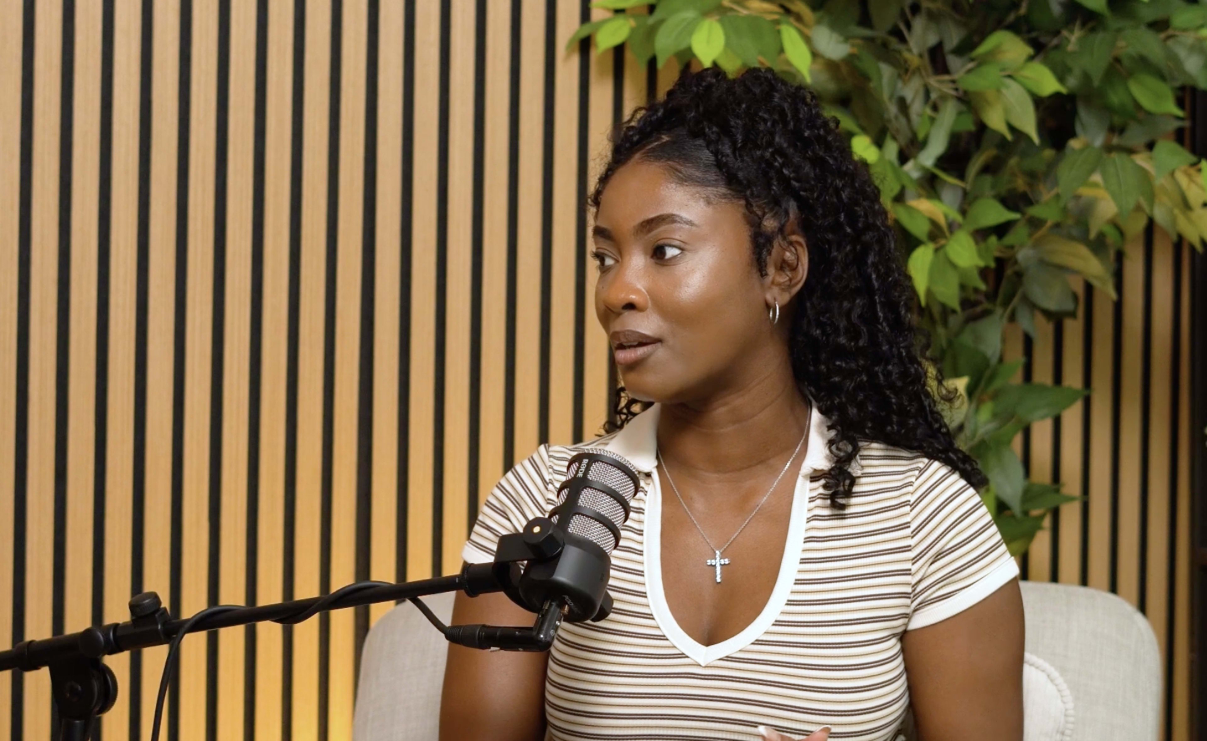 A woman with curly hair is speaking while sitting in front of a microphone, with a backdrop of vertical wooden panels and greenery.