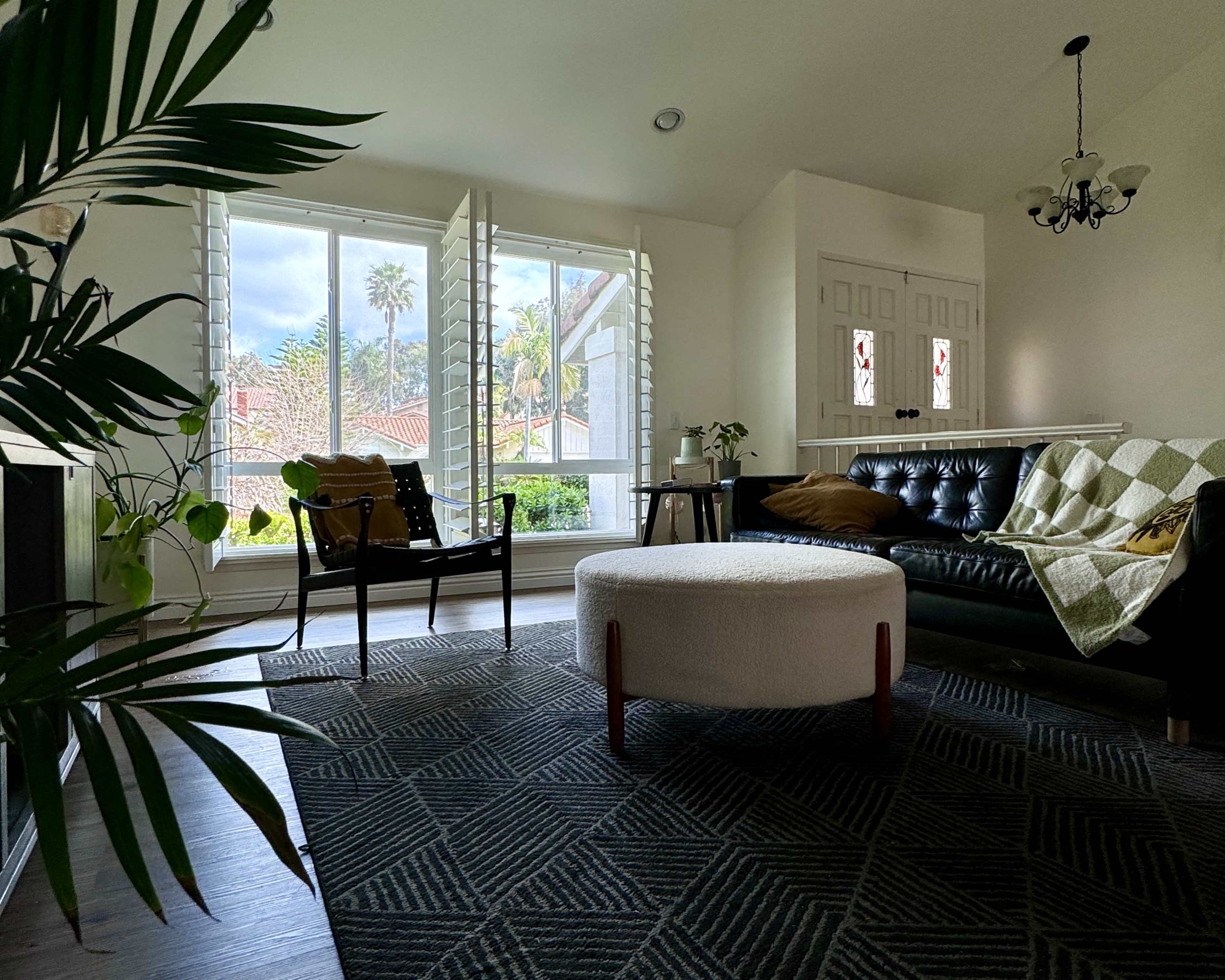 A modern living room with a black leather sofa, a round ottoman, large windows with shutters, and a patterned rug.