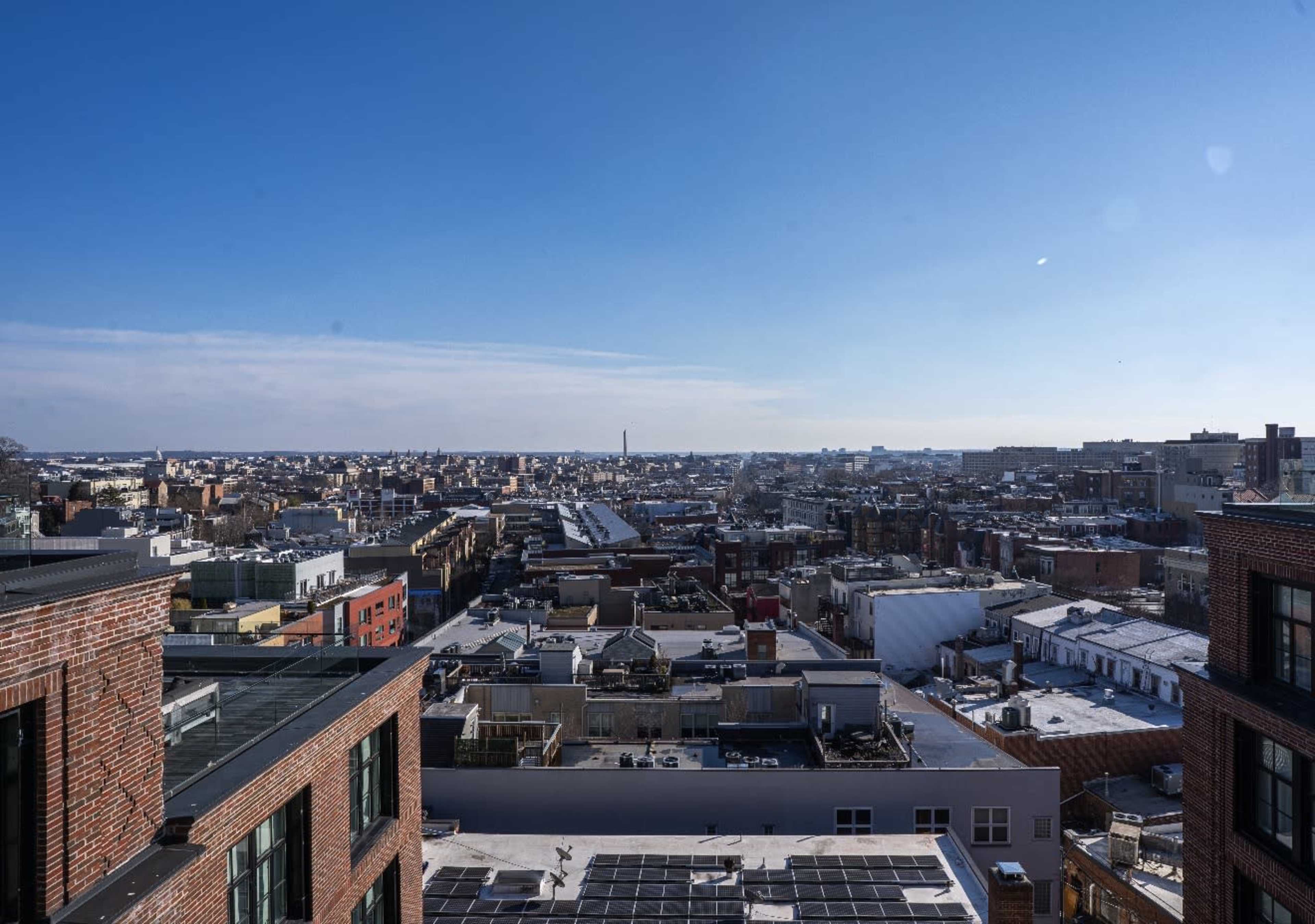 The image shows a panoramic view of a cityscape with a mix of buildings, rooftops, and distant landmarks under a clear blue sky.
