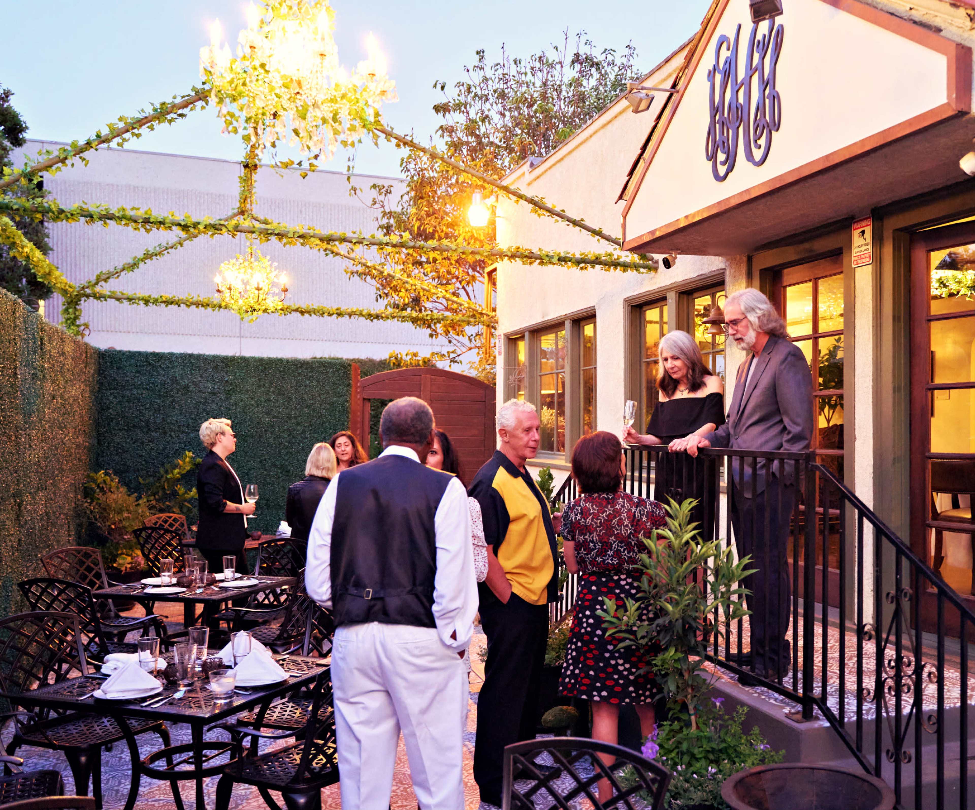 A group of people mingles on a patio adorned with string lights and greenery outside a restaurant.