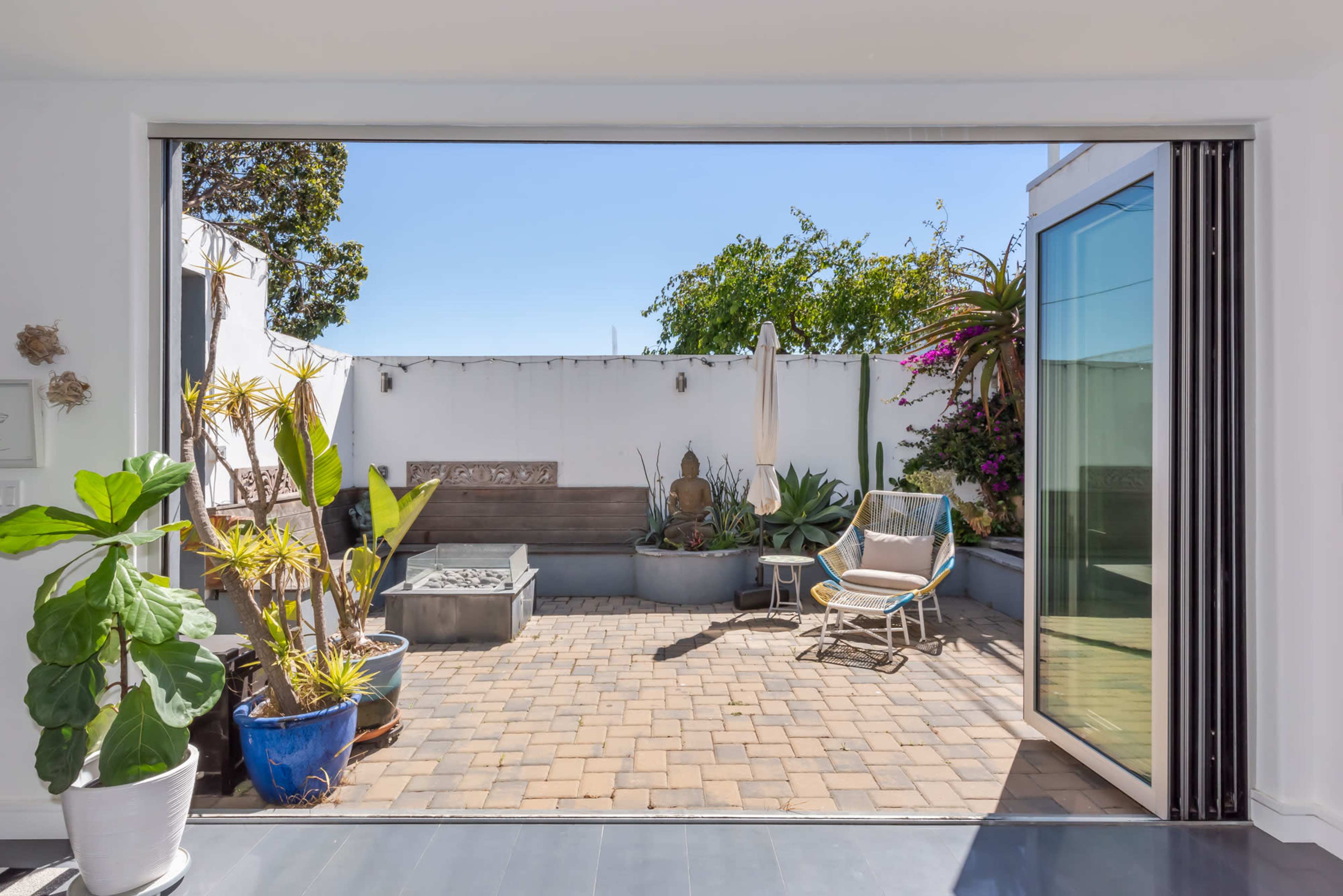A spacious patio with pavers features various plants, a lounge chair, and a small water fountain, enclosed by white walls and greenery.