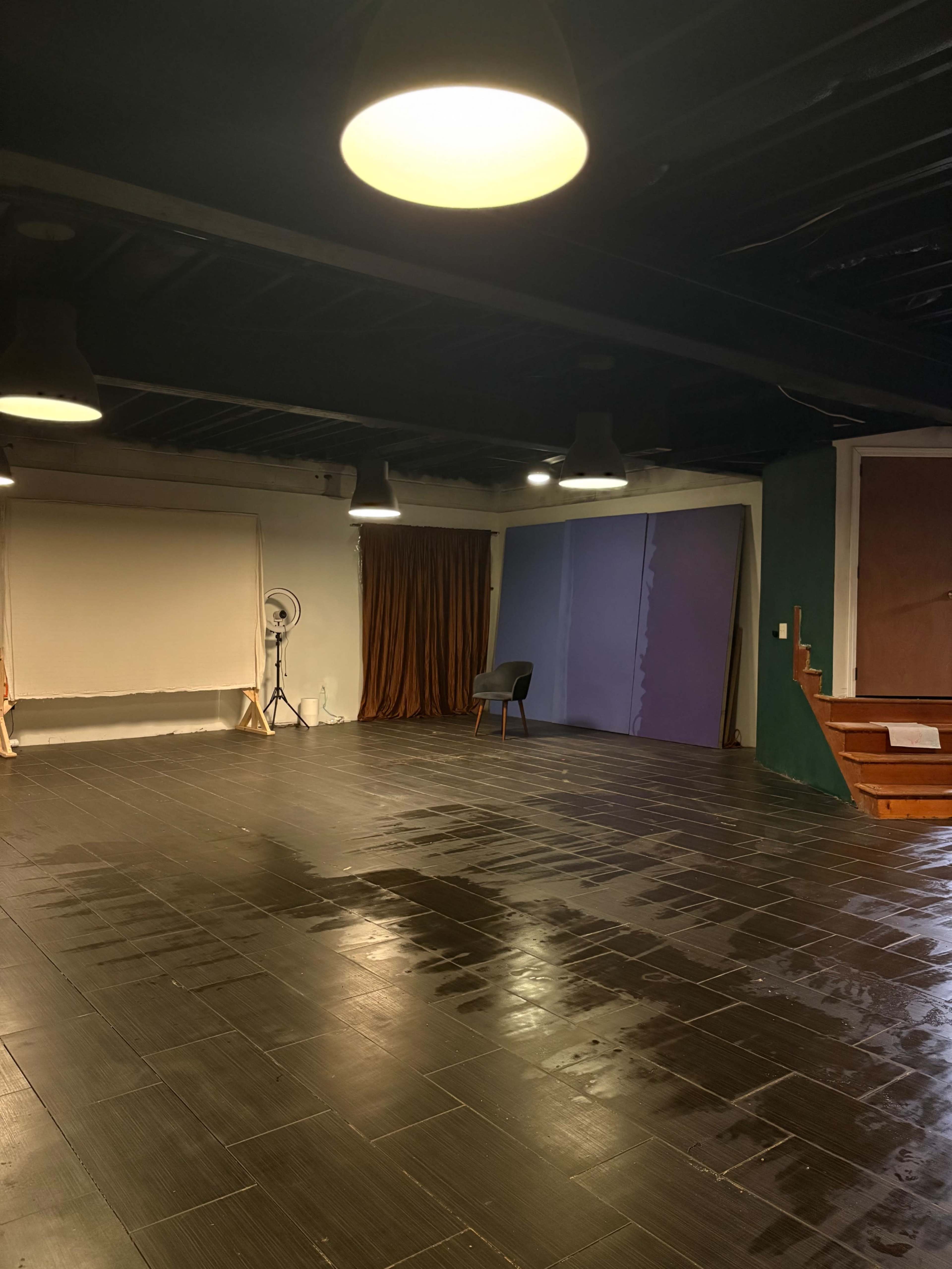 The image shows a spacious, dimly lit studio with a wooden floor, featuring a backdrop, a chair, and a curtain.