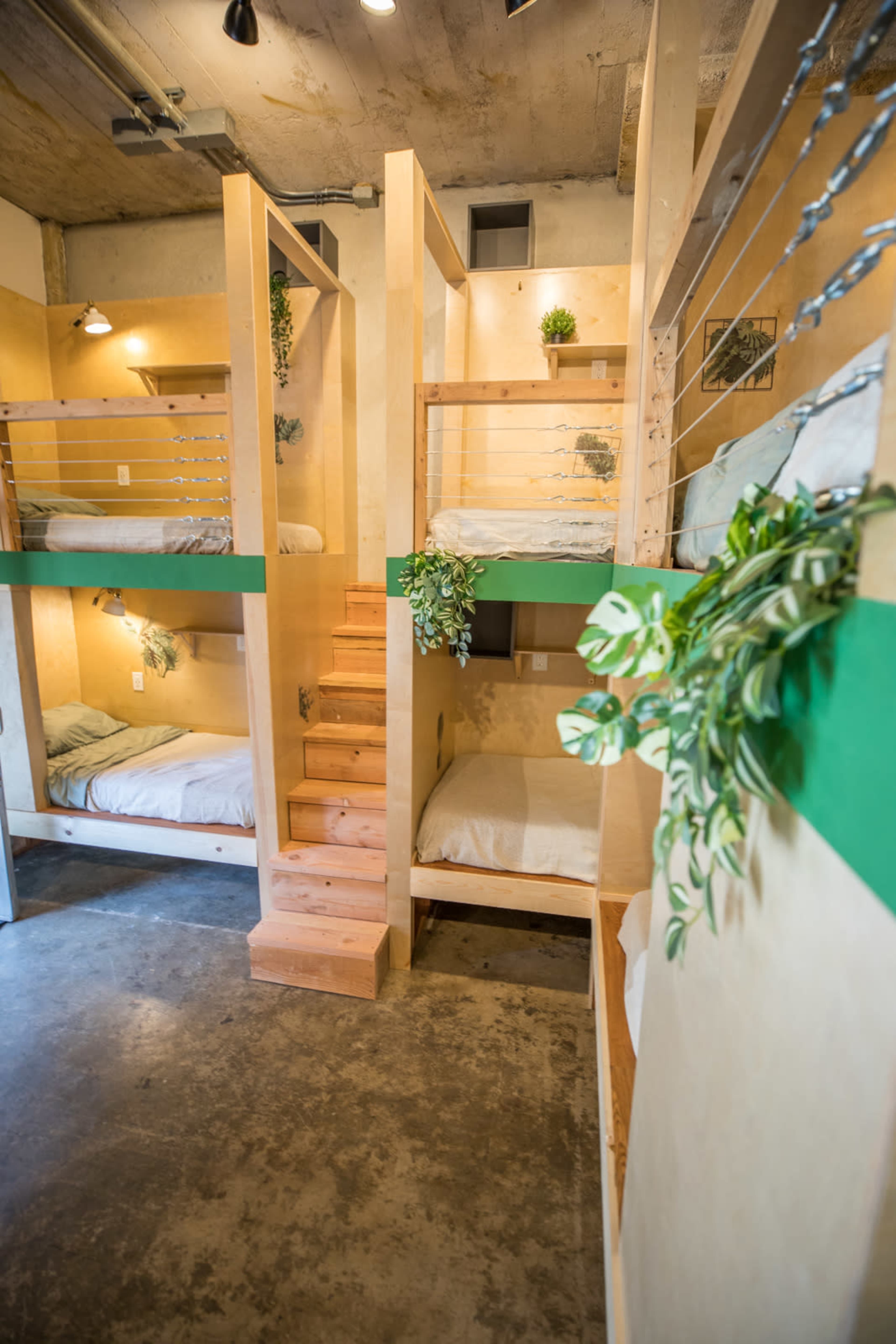 The image shows a modern multi-bunk sleeping area with wooden structures, green accents, and small plants integrated into the design.
