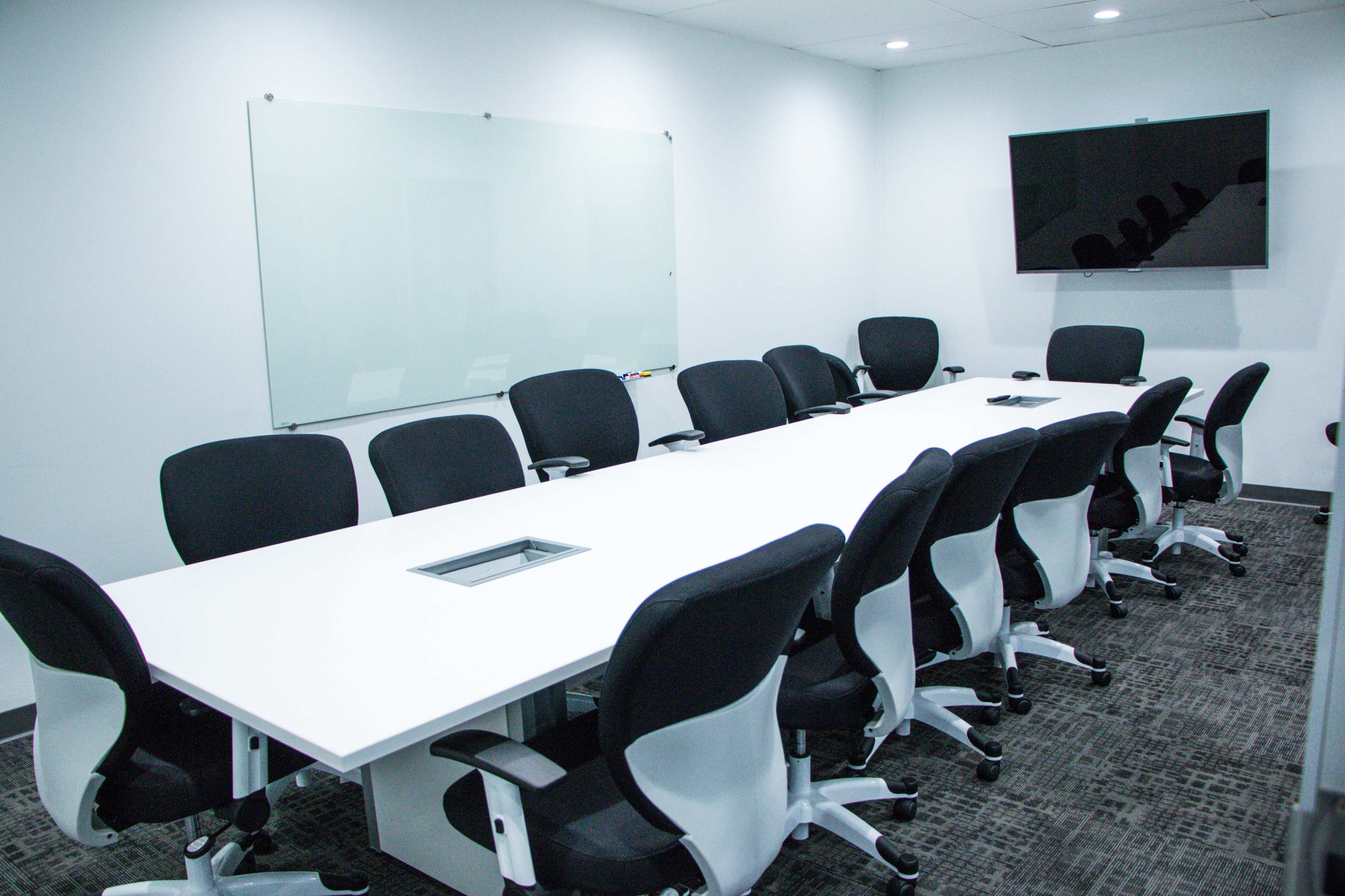 A large, modern conference room with a long white table surrounded by black office chairs, a wall-mounted TV, and a whiteboard.