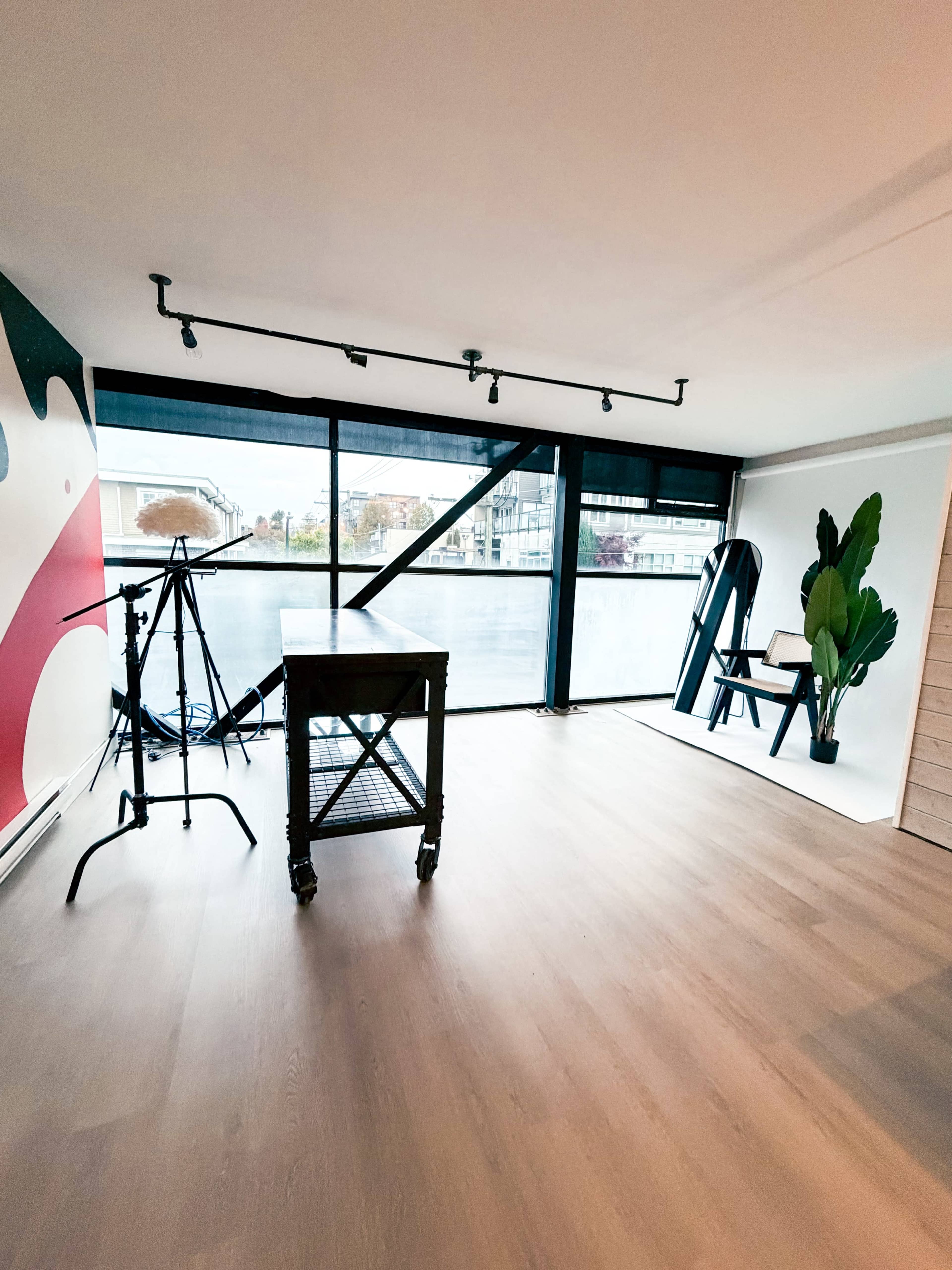 The image shows an empty room with large windows, a rolling cart, a photography tripod, and a plant next to a chair.