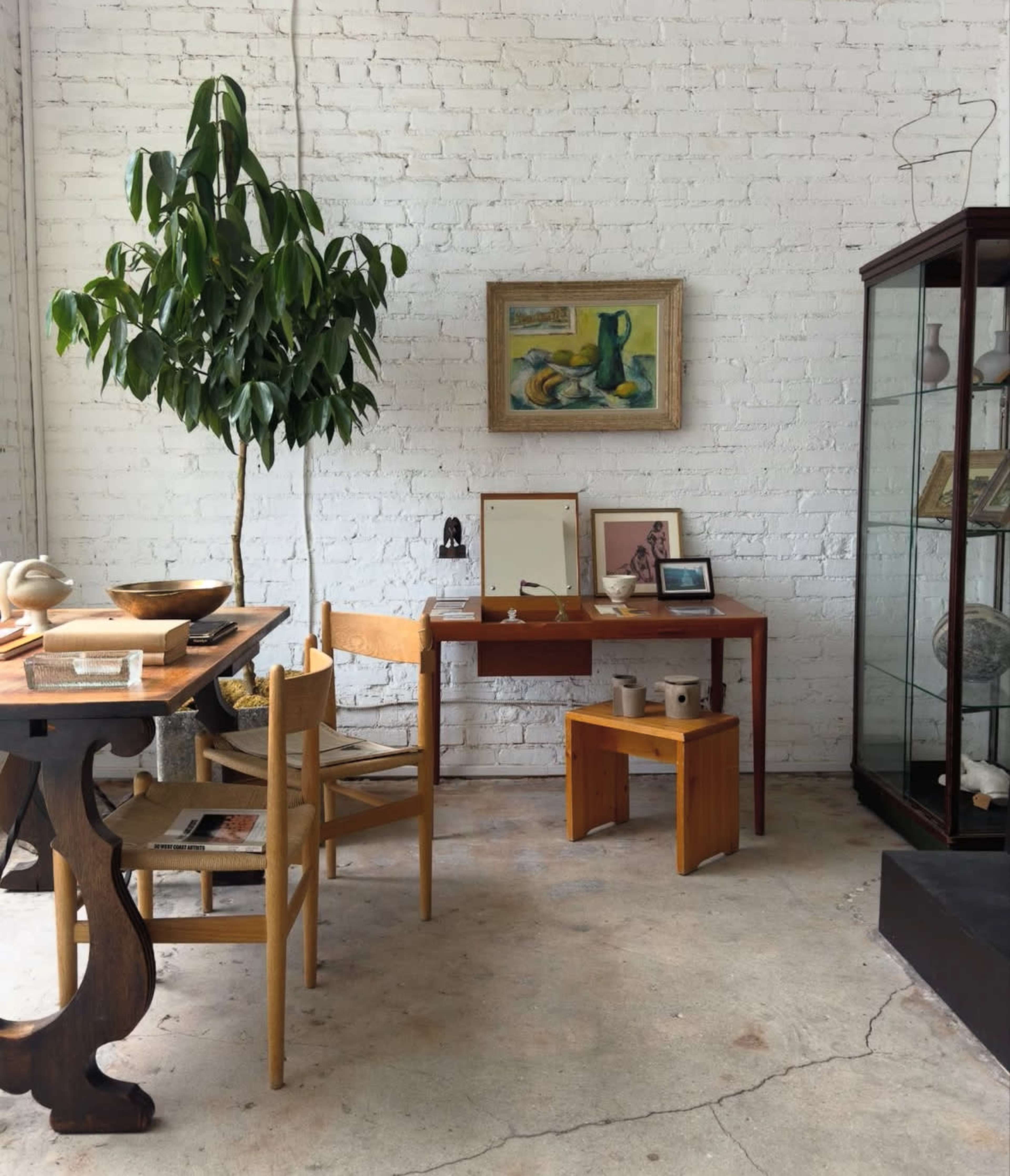 The image shows an interior space featuring a wooden table and chairs, a small stool, a wall-mounted artwork, a potted plant, and a glass display case.