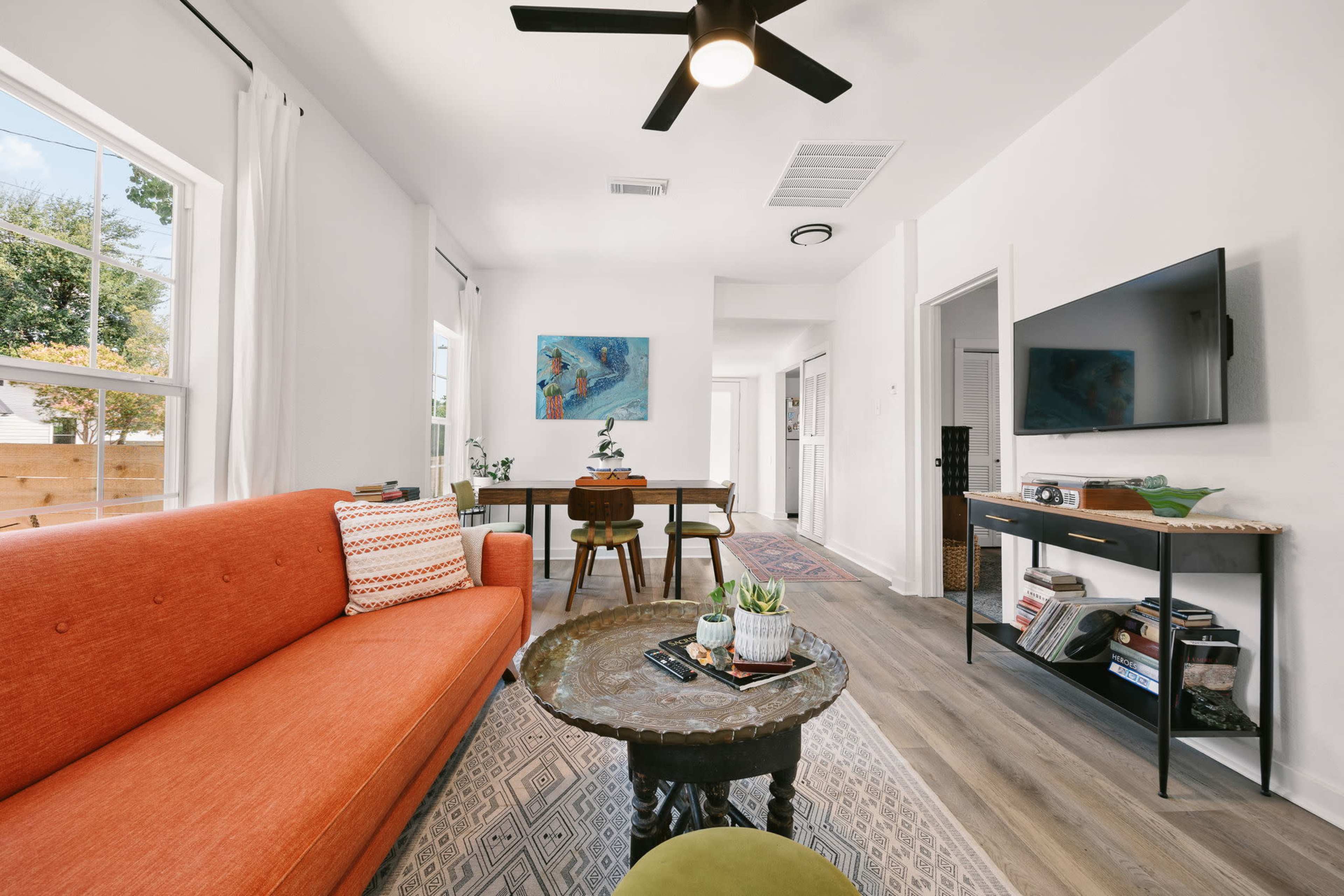 The image shows a modern living room featuring an orange sofa, a round coffee table with a plant, and a dining area in the background, complemented by a TV and a few decorative elements.