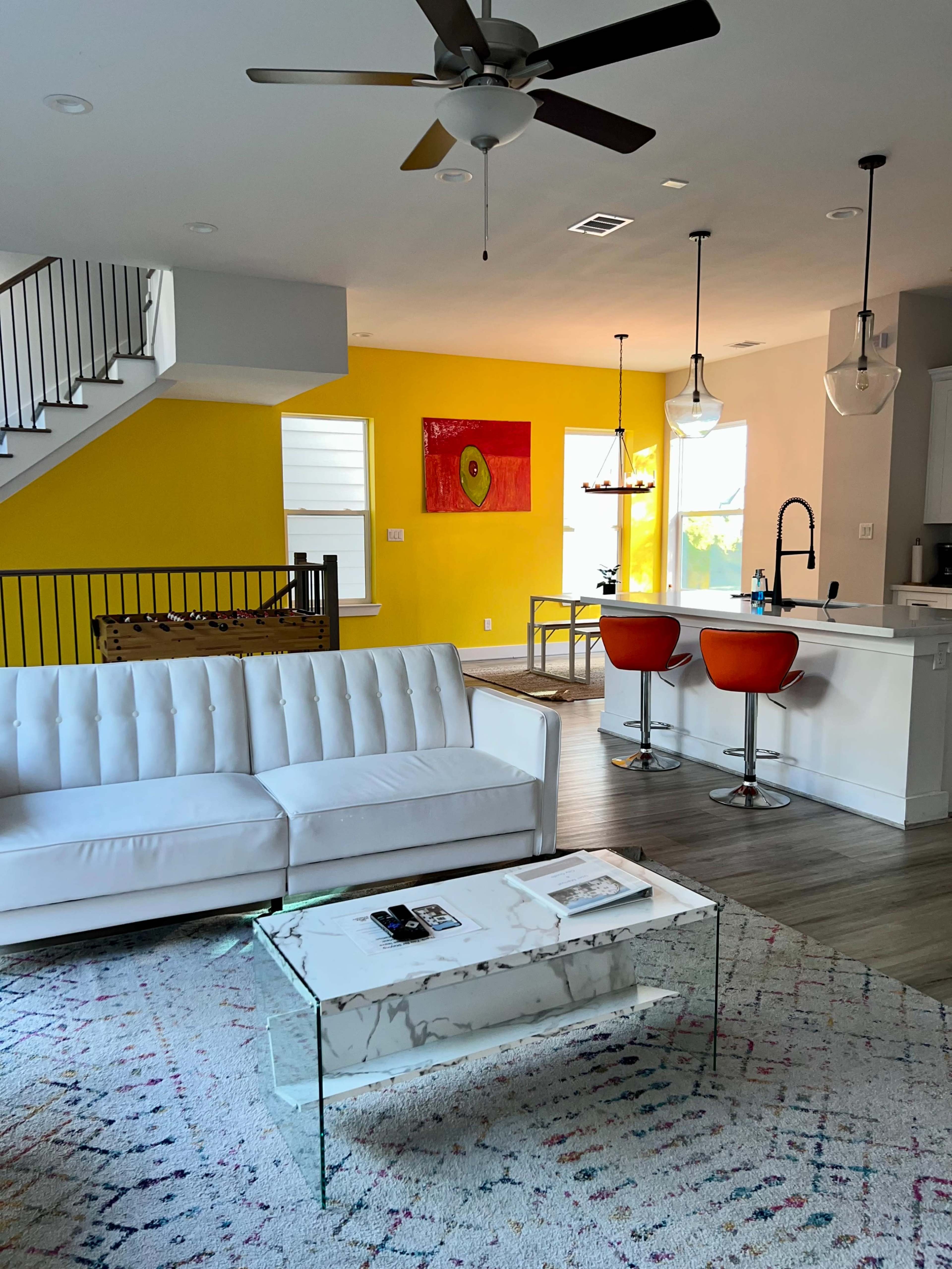 A modern living area with a white couch, a glass coffee table, and orange bar stools at a kitchen island, accented by a yellow wall and a colorful painting.