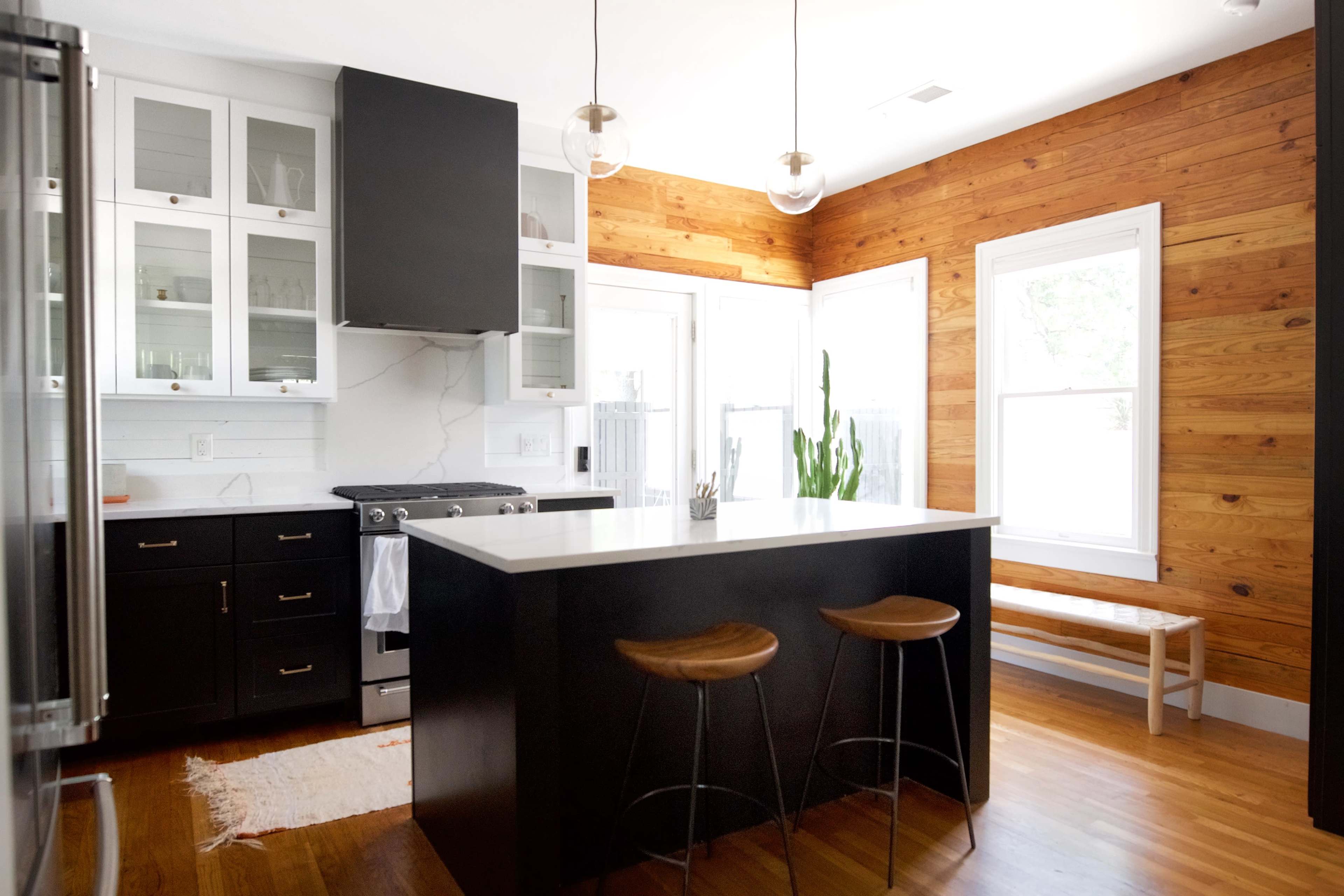 A modern kitchen with black cabinets, a white countertop, and wooden accents, featuring two stools at the island and a large window.