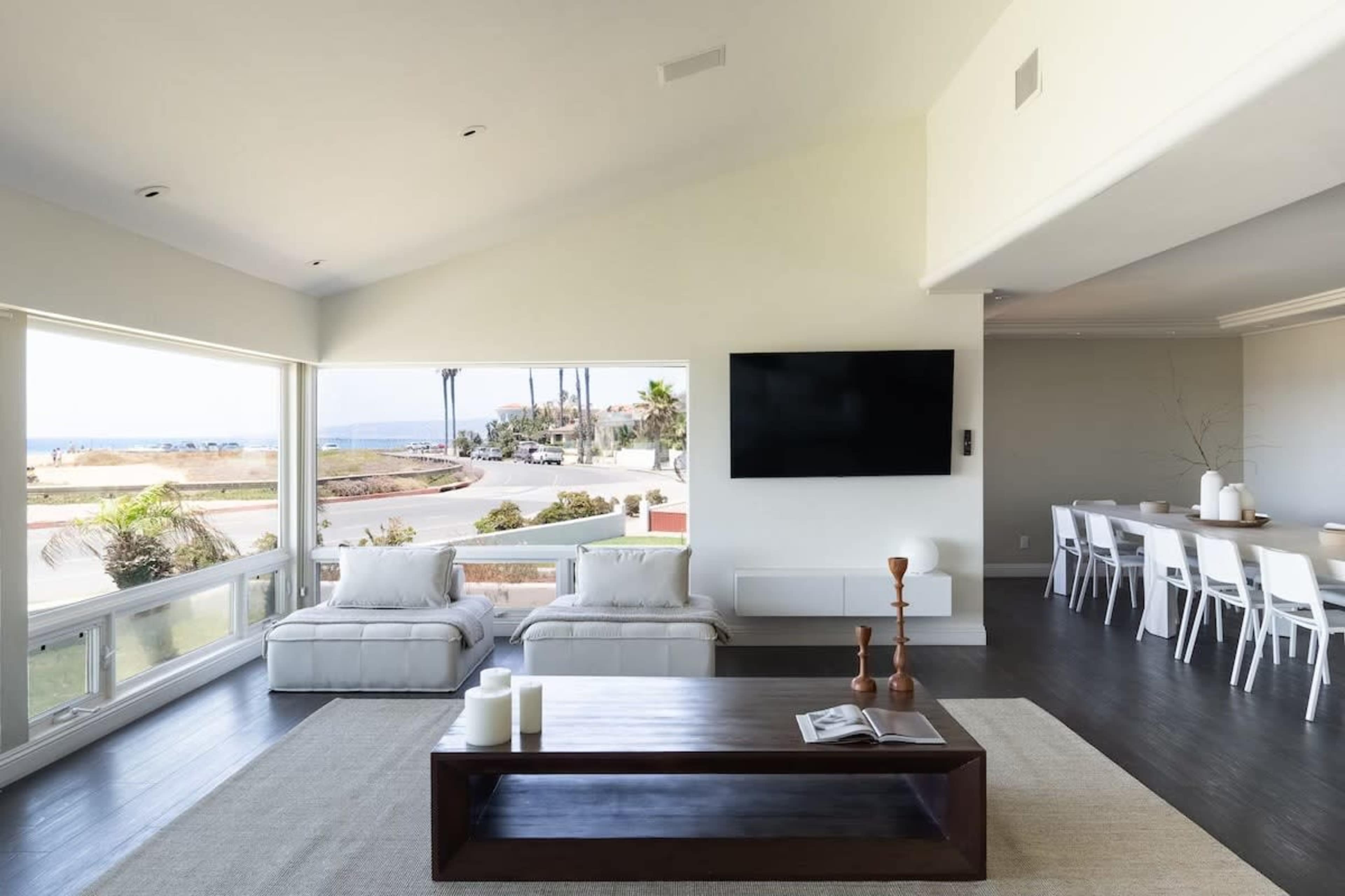 A spacious modern living room features two white sofas facing large windows with a view of the ocean, alongside a wooden coffee table and a dining area in the background.