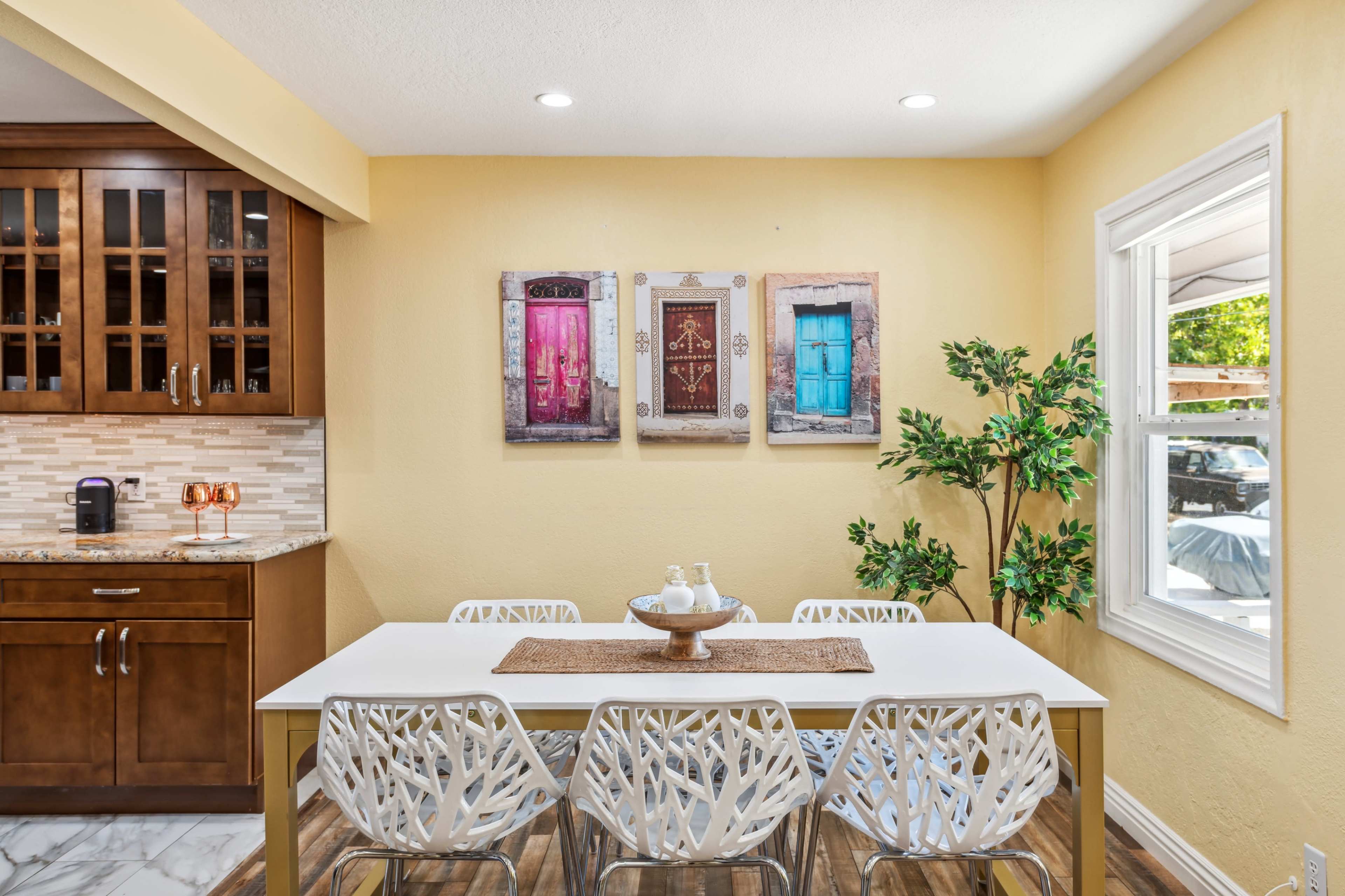 A dining area features a white table surrounded by modern chairs, with colorful framed doors on the wall and a potted plant in the corner.