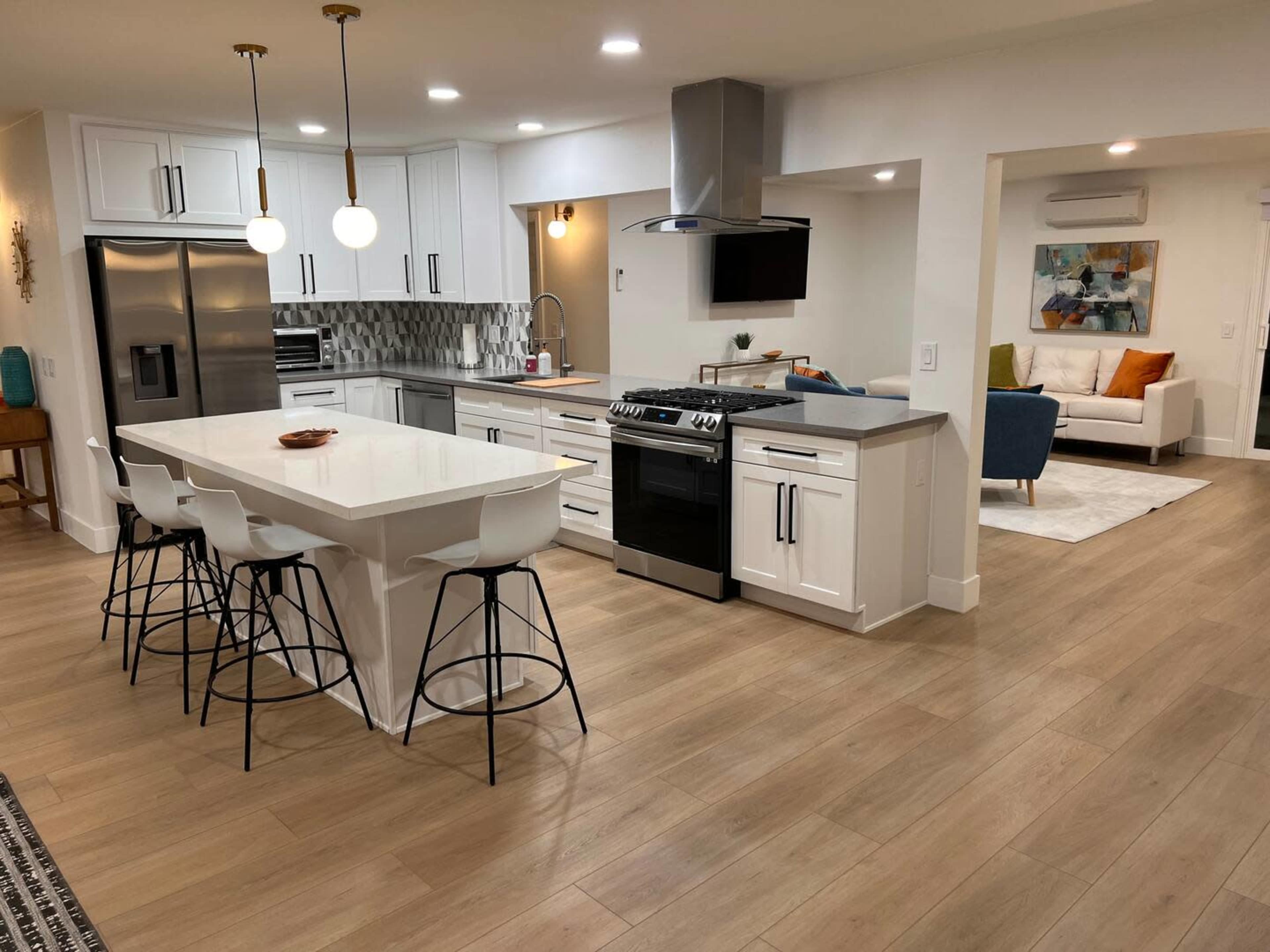 A modern kitchen with white cabinetry, a large island with seating, stainless steel appliances, and an adjoining living area.
