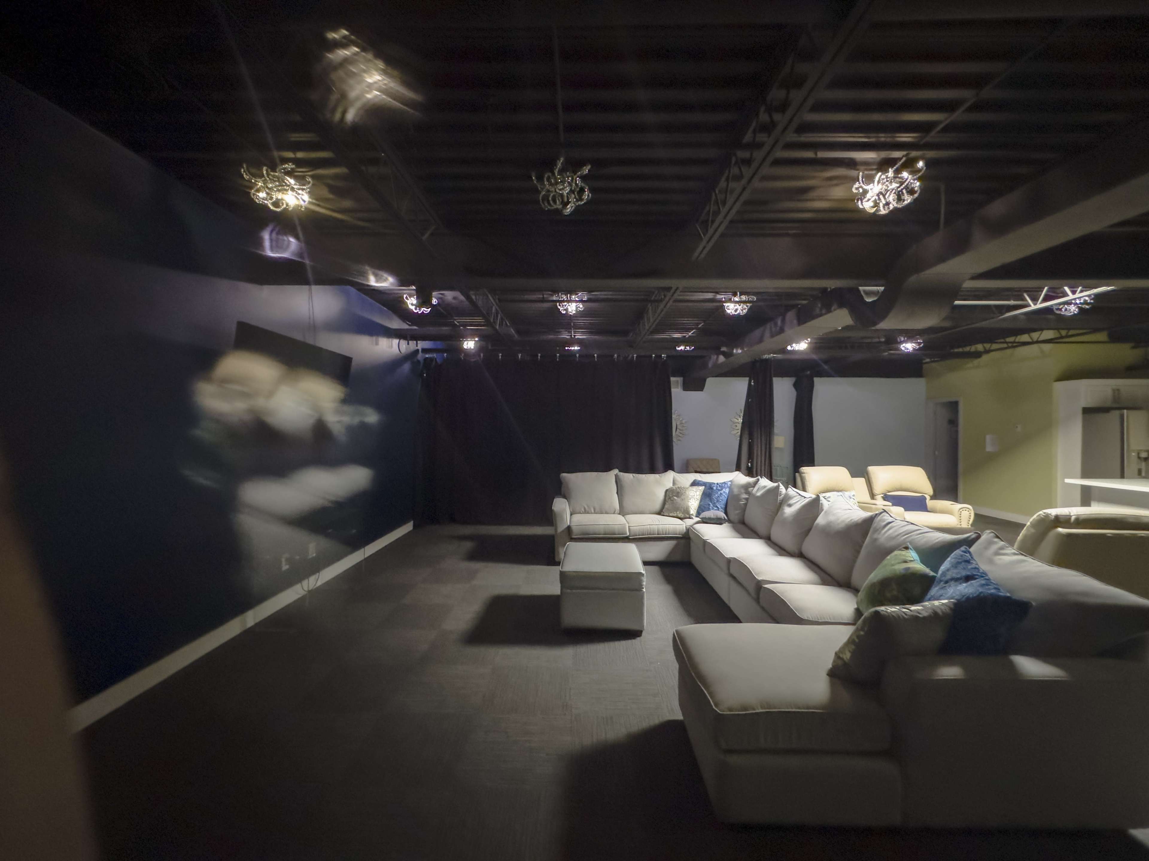 A spacious room with a large gray sectional sofa, a dark ceiling, and dim lighting, featuring a backdrop of black curtains.