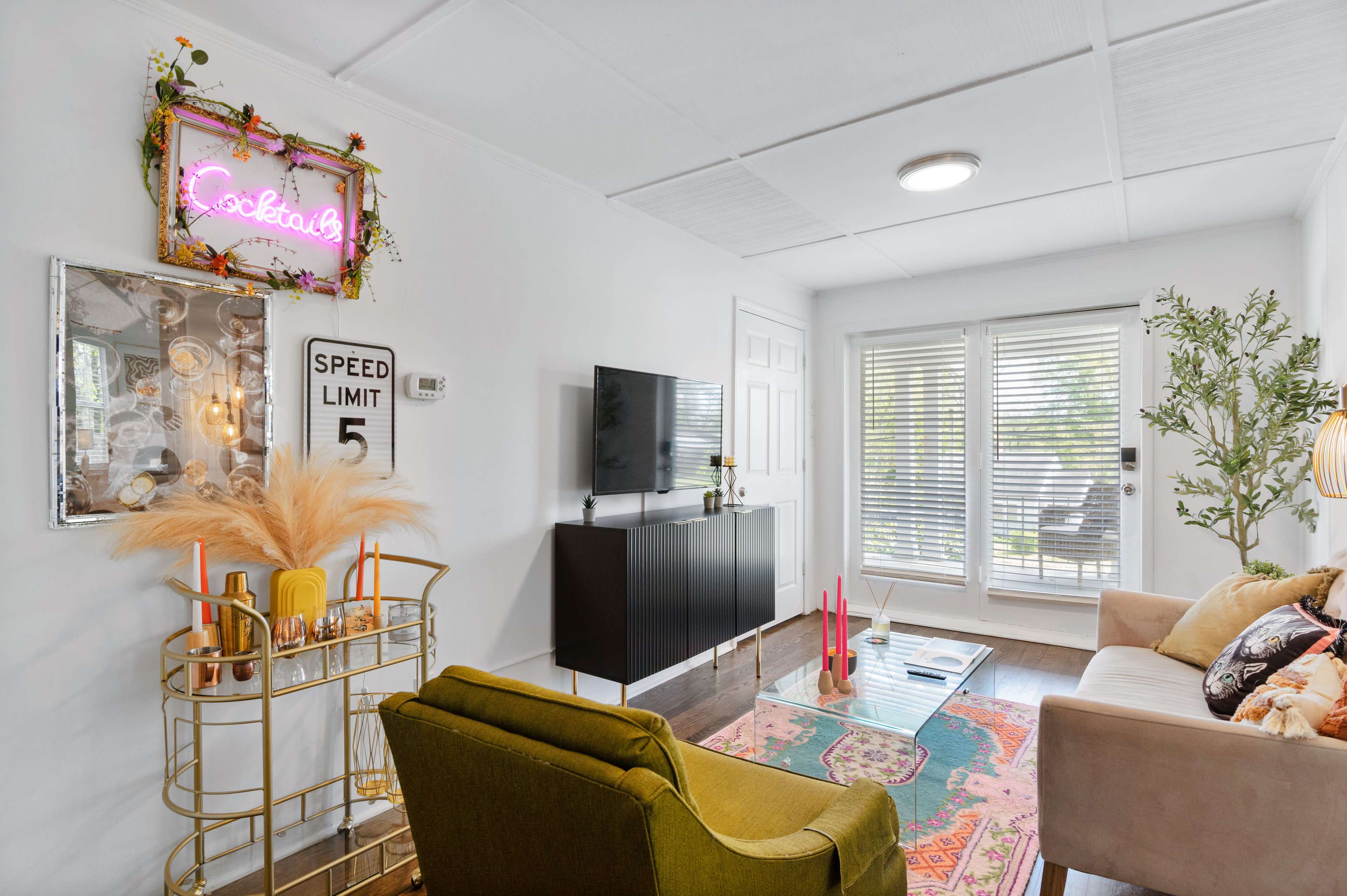 A cozy living room featuring a green armchair, a sleek black TV stand with a television, and a vibrant rug, with large sliding glass doors leading to an outdoor area.