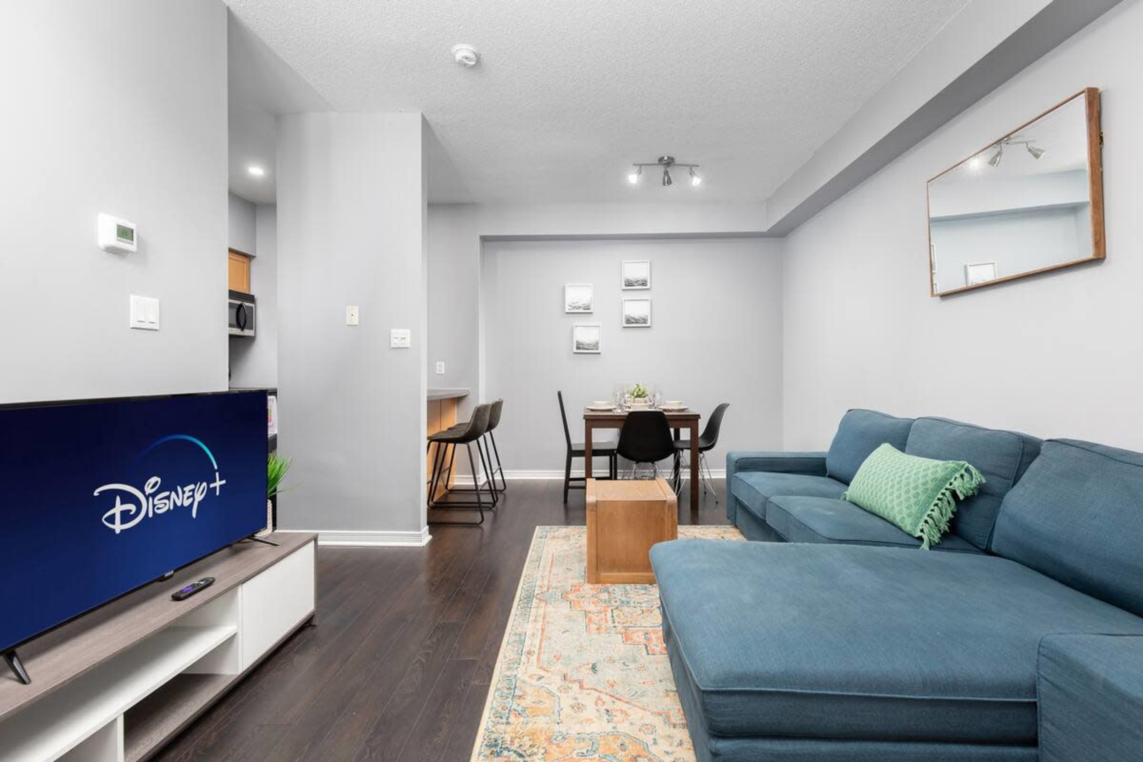 The image shows a modern living room with a blue sofa, a wooden coffee table, a television displaying the Disney+ logo, and a dining area in the background.