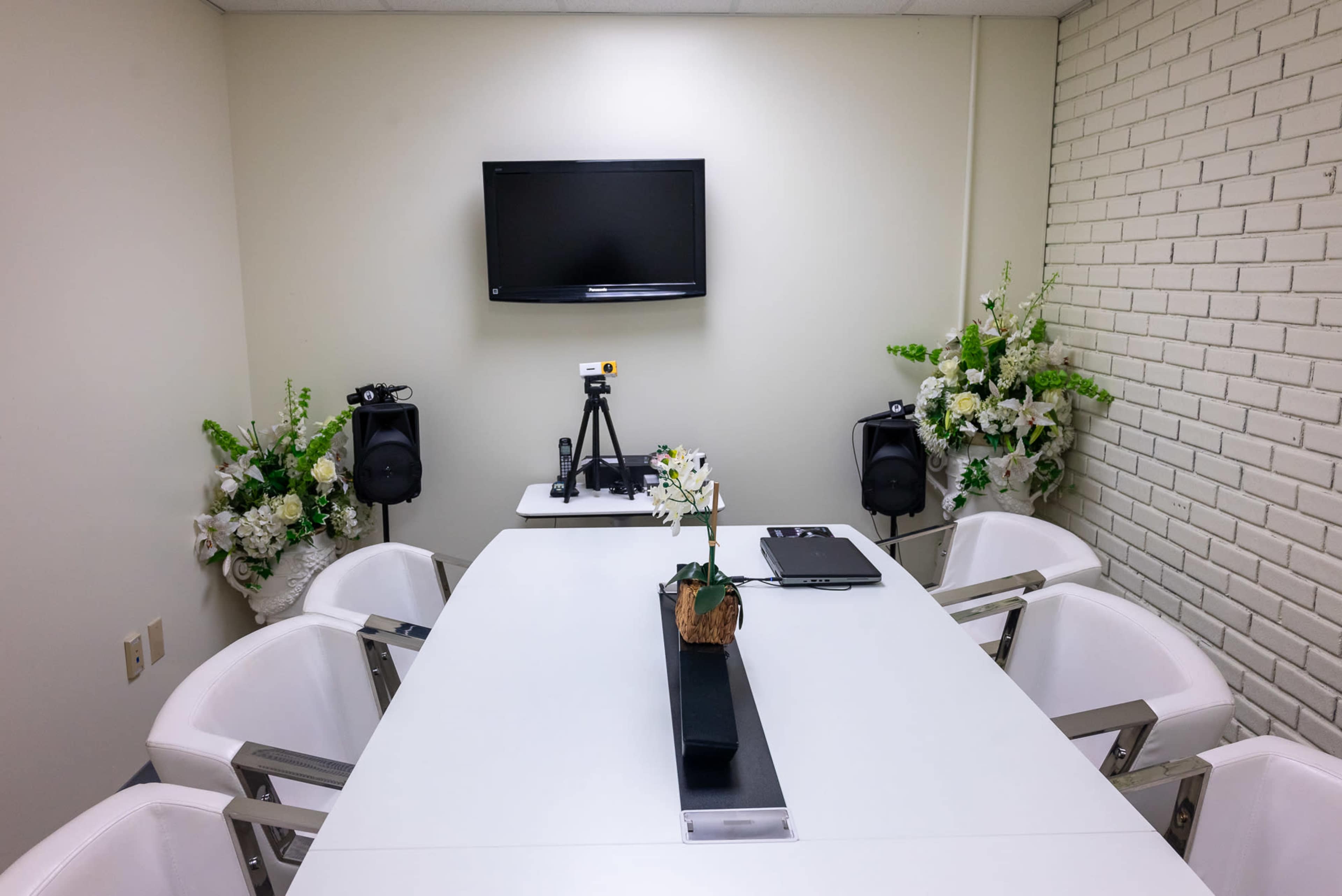 A meeting room features a long white table surrounded by six white chairs, with a television mounted on the wall and decorative flower arrangements in the corners.