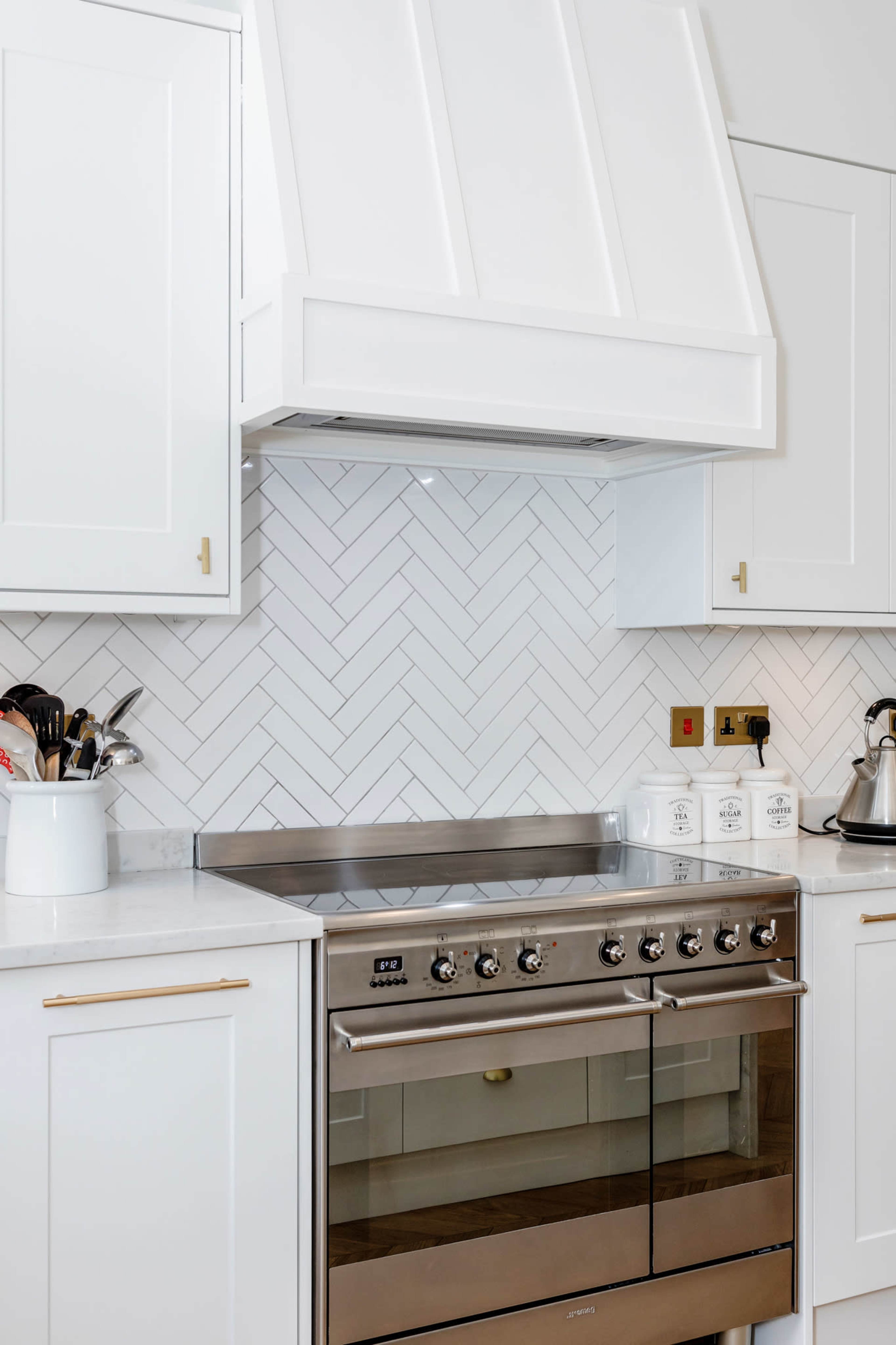 The image shows a modern kitchen featuring a stainless steel range oven beneath a white herringbone tile backsplash, with white cabinetry and cooking utensils displayed.