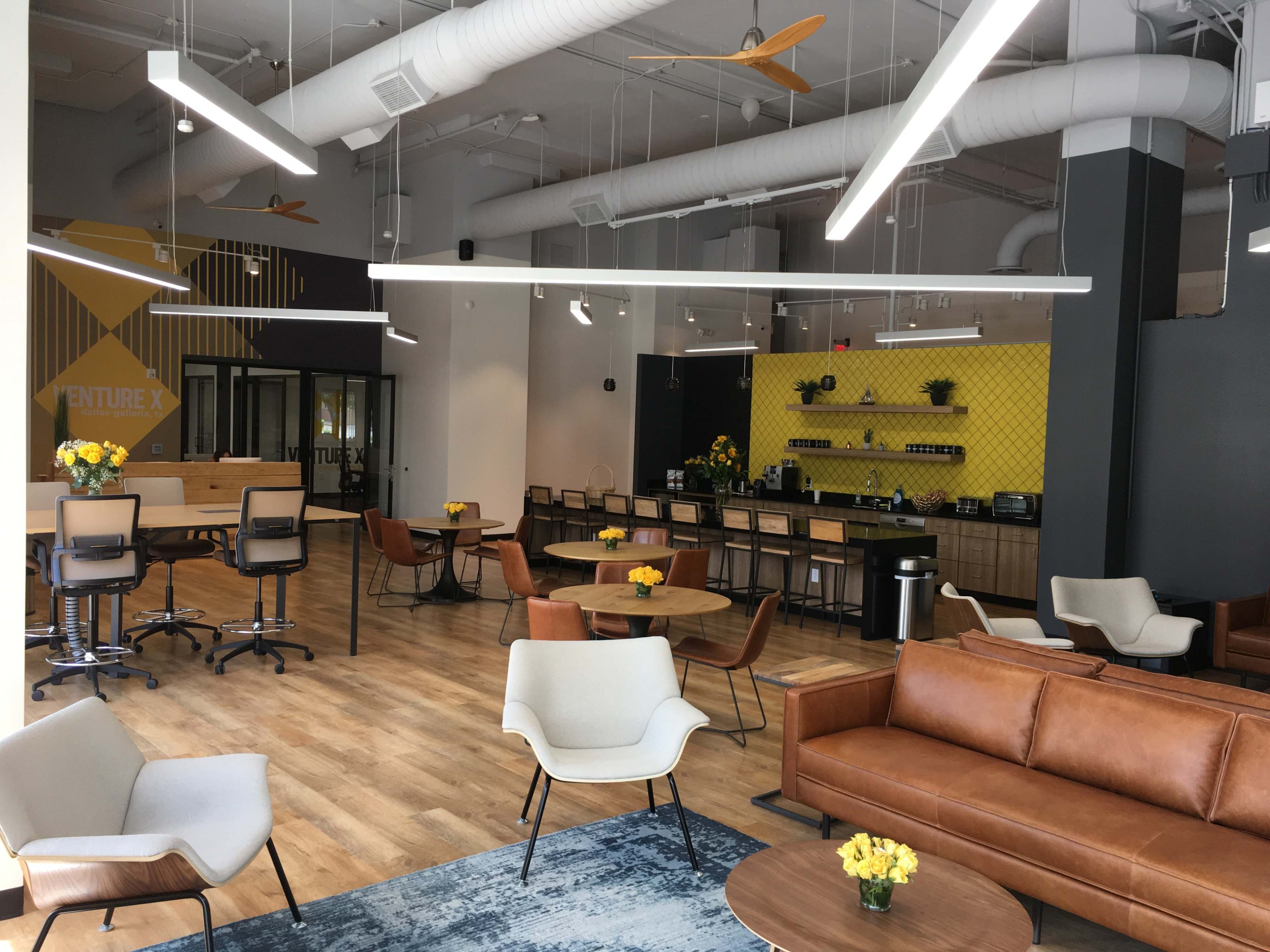A modern coworking space with various seating areas, yellow flowers on tables, and a kitchenette in the background.