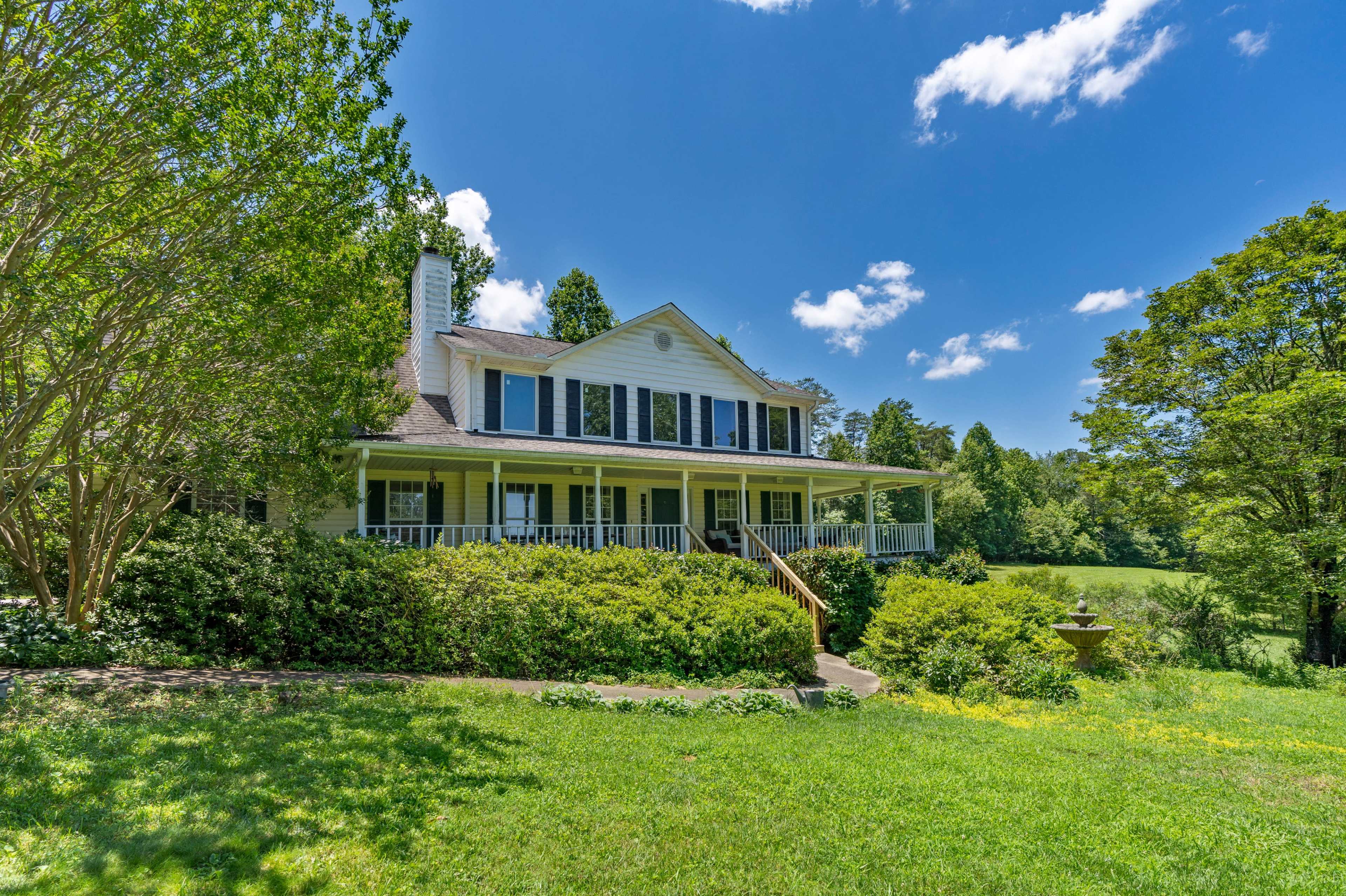A large white house with a porch and blue shutters is surrounded by greenery under a clear blue sky.