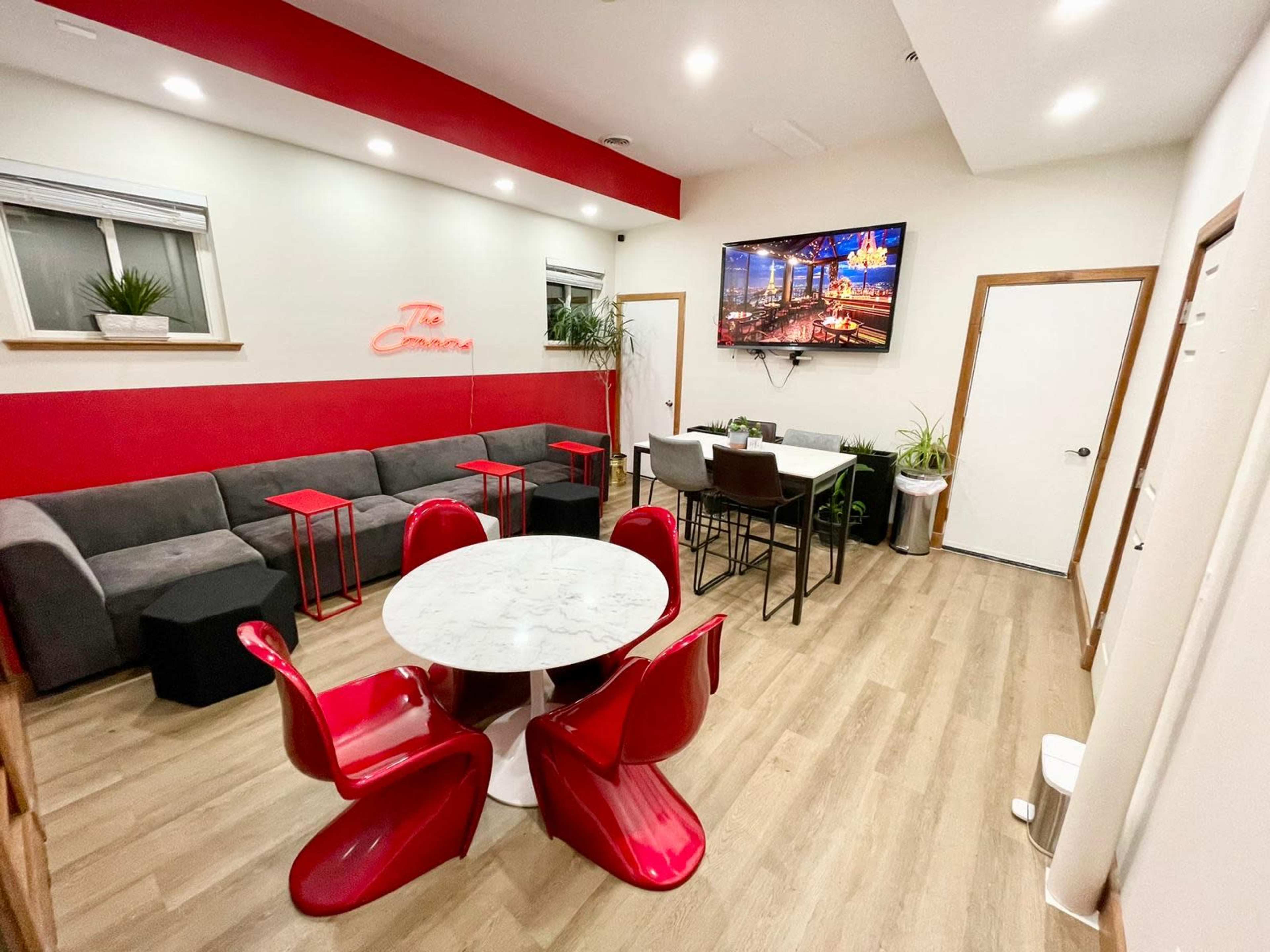 A modern, minimalist lounge area featuring a gray sofa, red accent walls, a marble table surrounded by red chairs, and a large TV mounted on the wall.