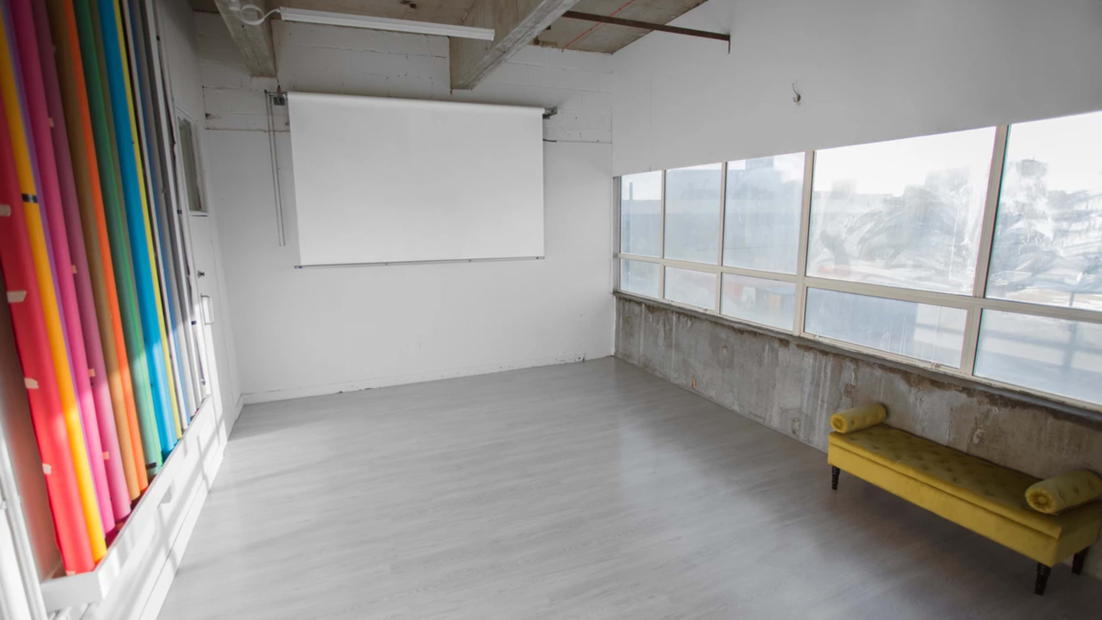 A bright, minimalist interior space features a large window, a blank projector screen, and a yellow upholstered bench.