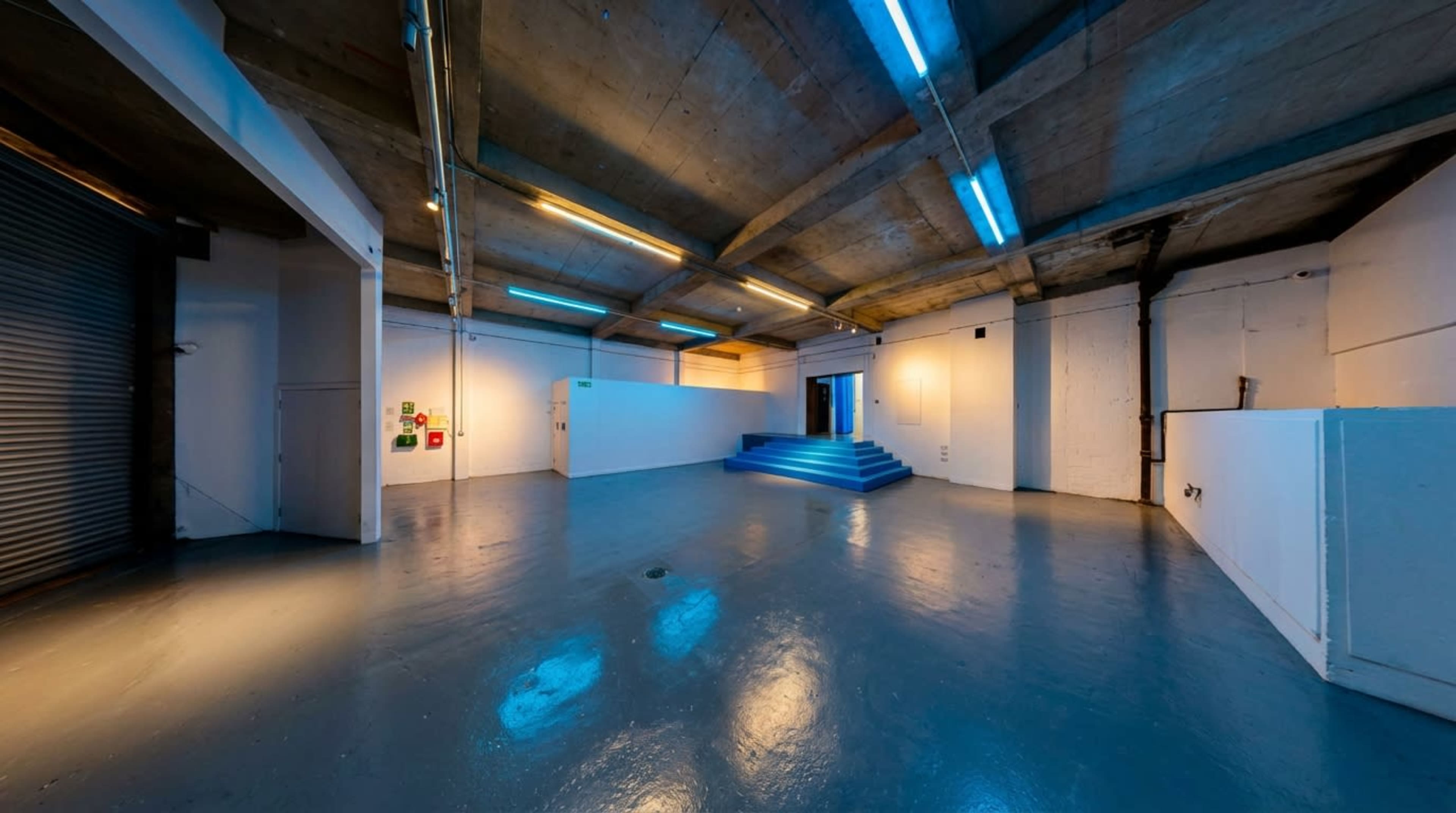 The image shows a large, empty industrial space with concrete walls and a polished floor, featuring blue lighting and a staircase leading to a doorway.