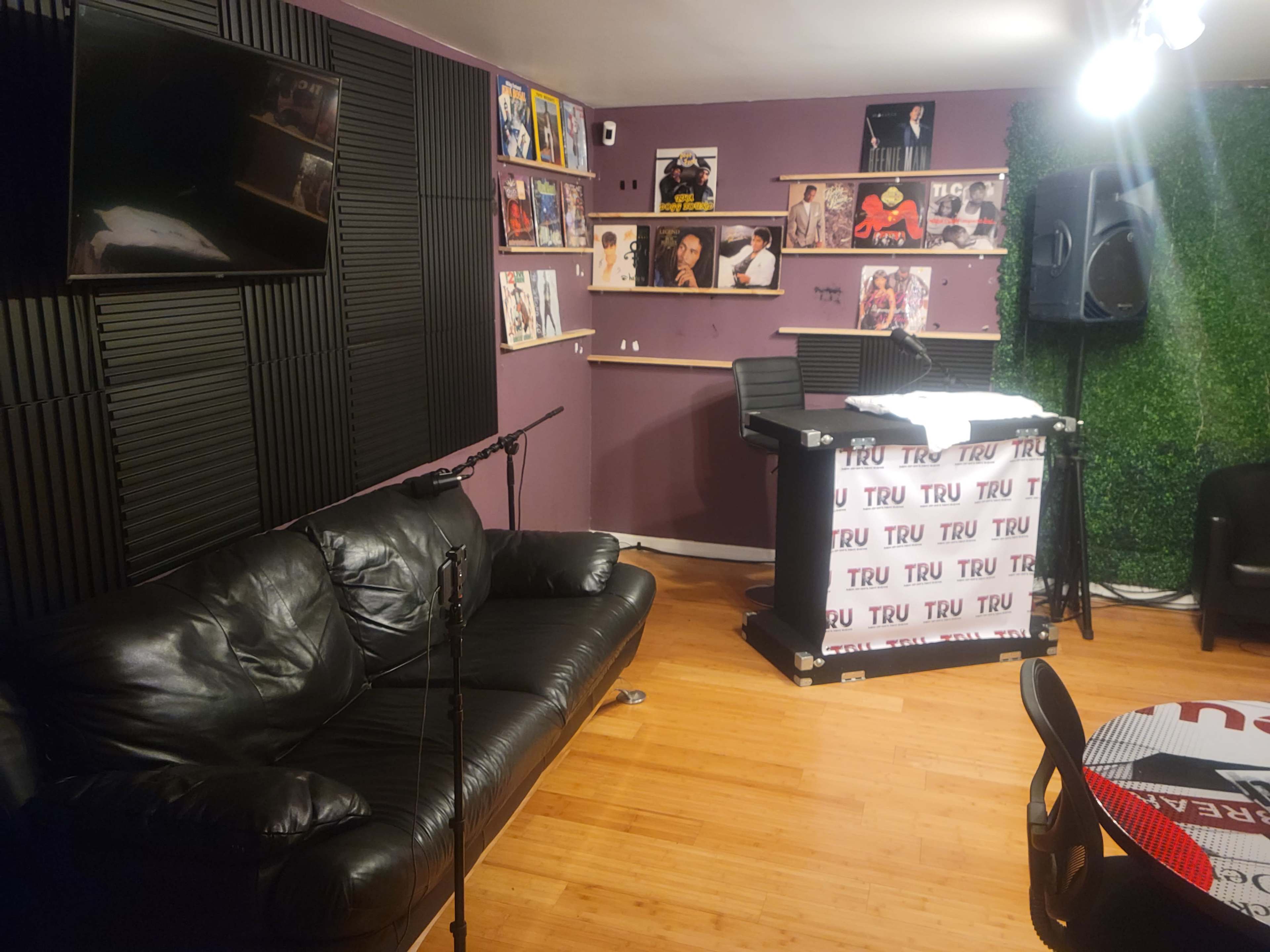 A recording studio with a black leather couch, a microphone, a desk set up for recording, and wall-mounted shelves displaying various artwork and albums.