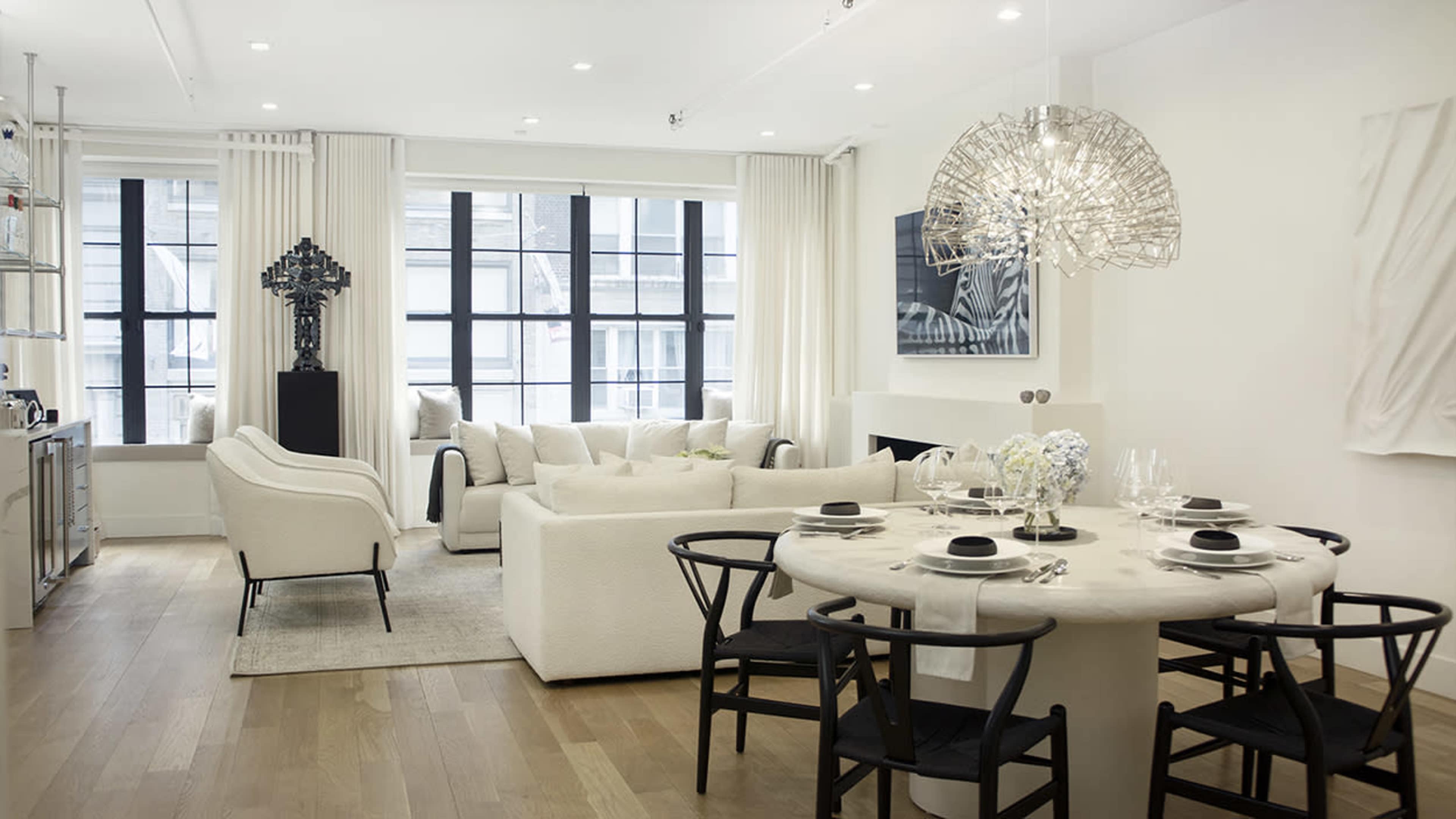 The image shows a modern interior living space featuring a round dining table with black chairs, a beige sofa, and large windows with sheer curtains.
