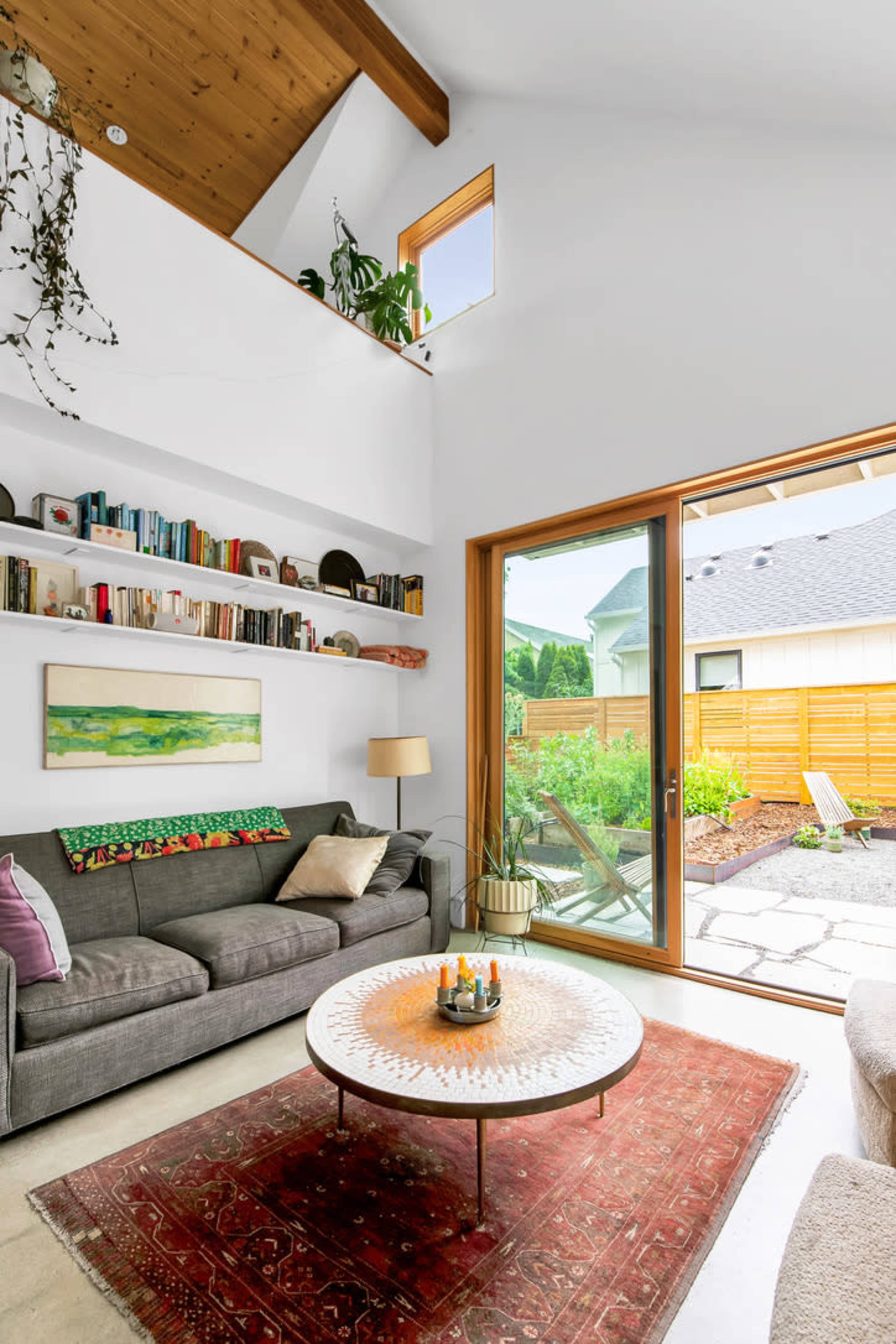 A living room features a gray sofa, a circular coffee table with candles, and large windows that provide a view of a landscaped outdoor area.