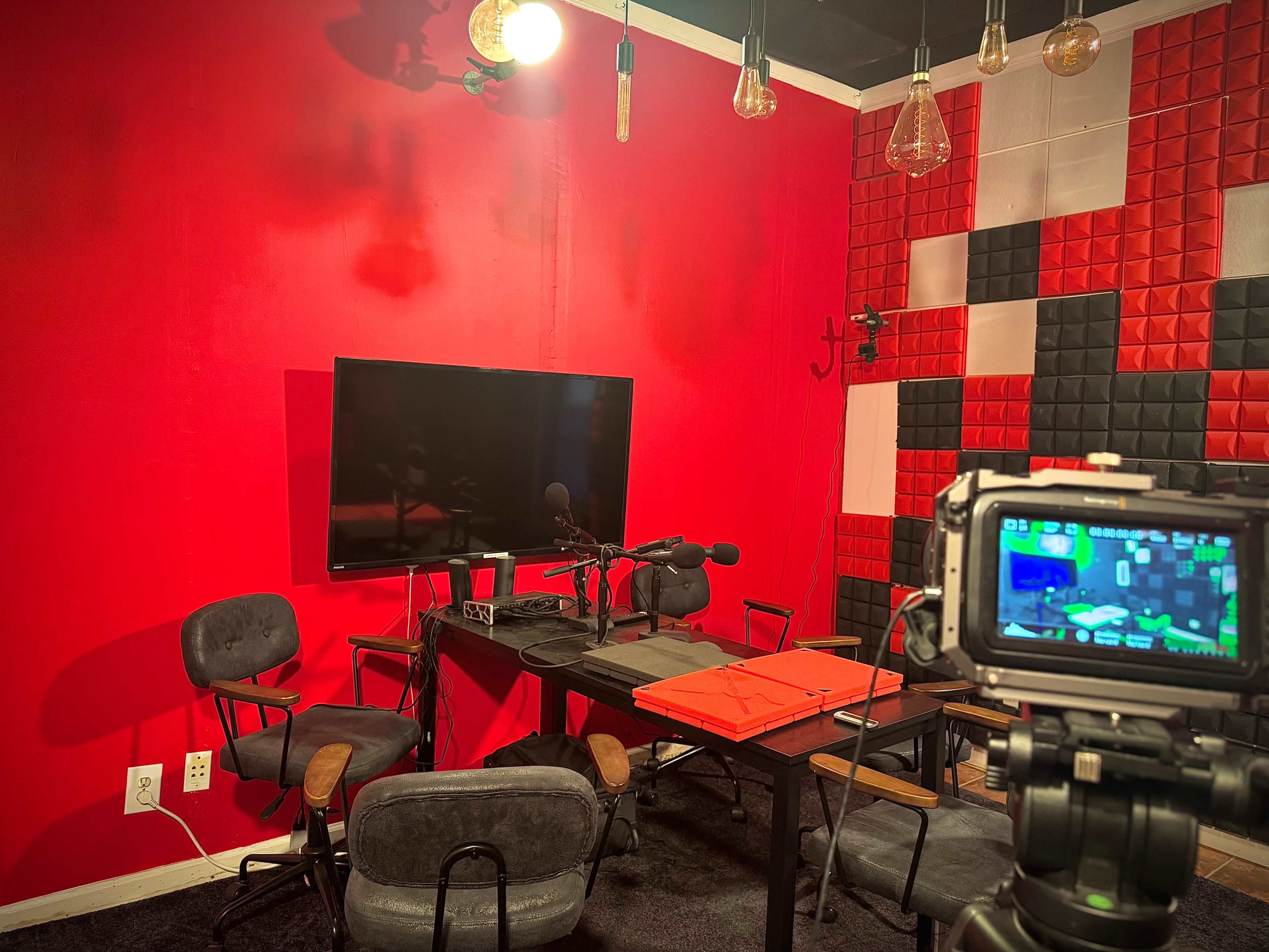 The image shows a small recording studio with a large TV screen, a table with microphones, and several chairs, all set against a vibrant red wall.