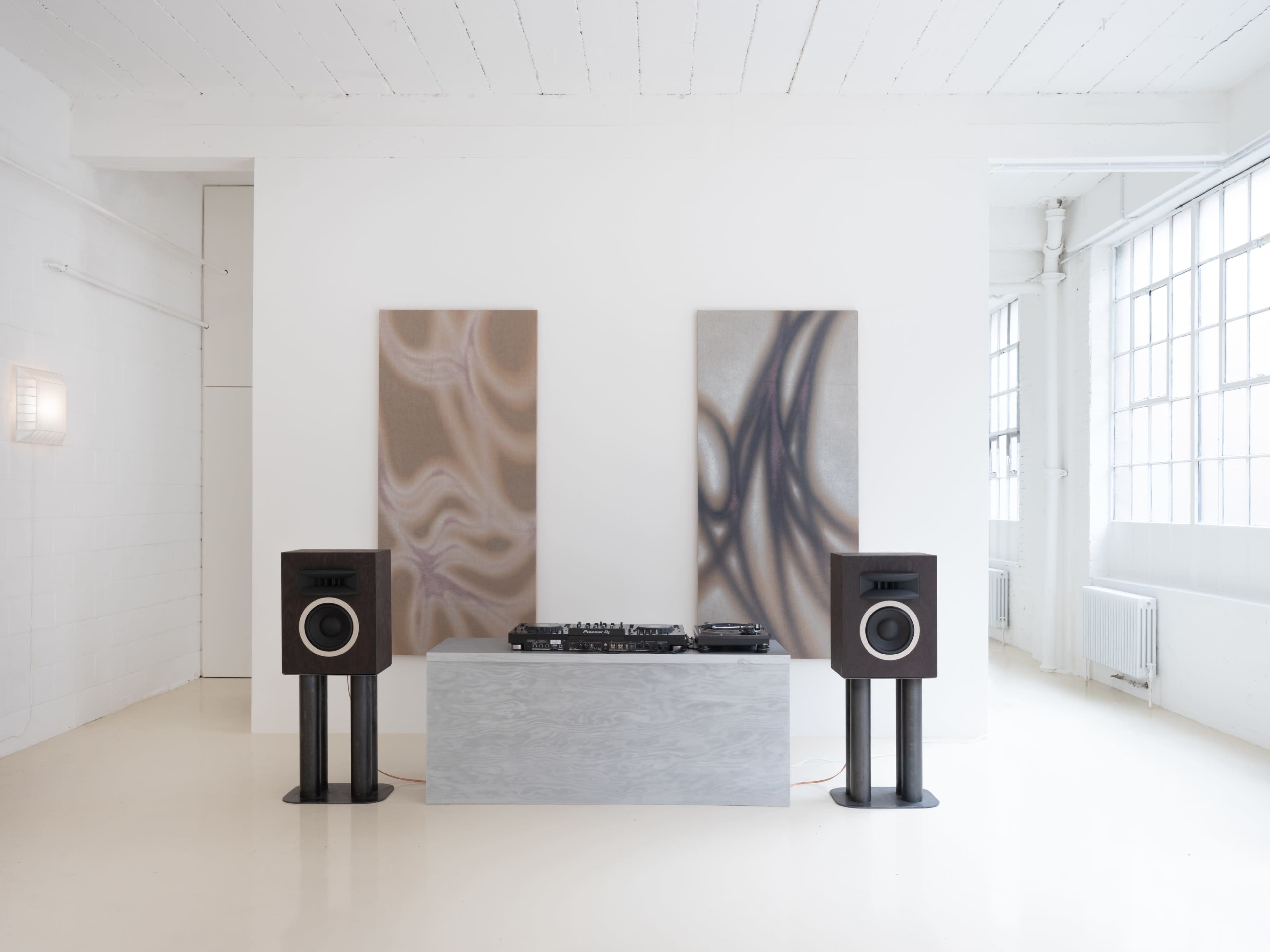 The image features a minimalist room with two abstract paintings on the walls, a DJ setup on a light-colored table, and speakers placed on stands on either side of the table.