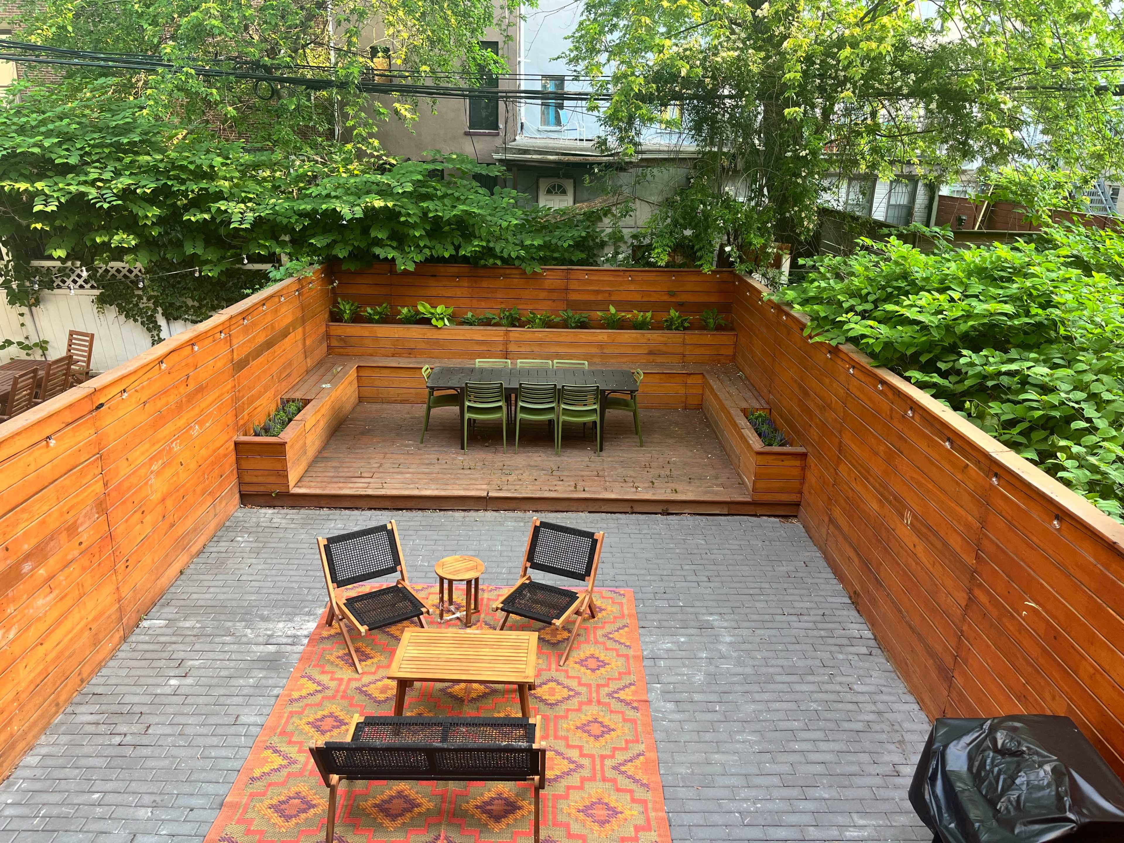 The image shows a landscaped outdoor patio area with wooden seating, a dining table, and greenery surrounding the space.