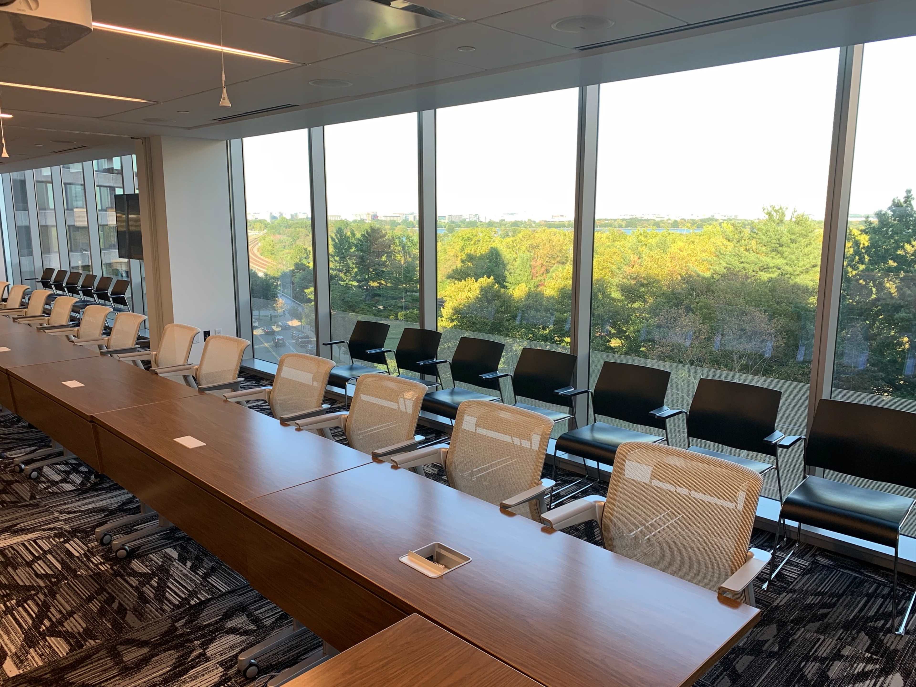 A modern conference room features a long table with multiple chairs set against large windows showcasing a view of greenery outside.