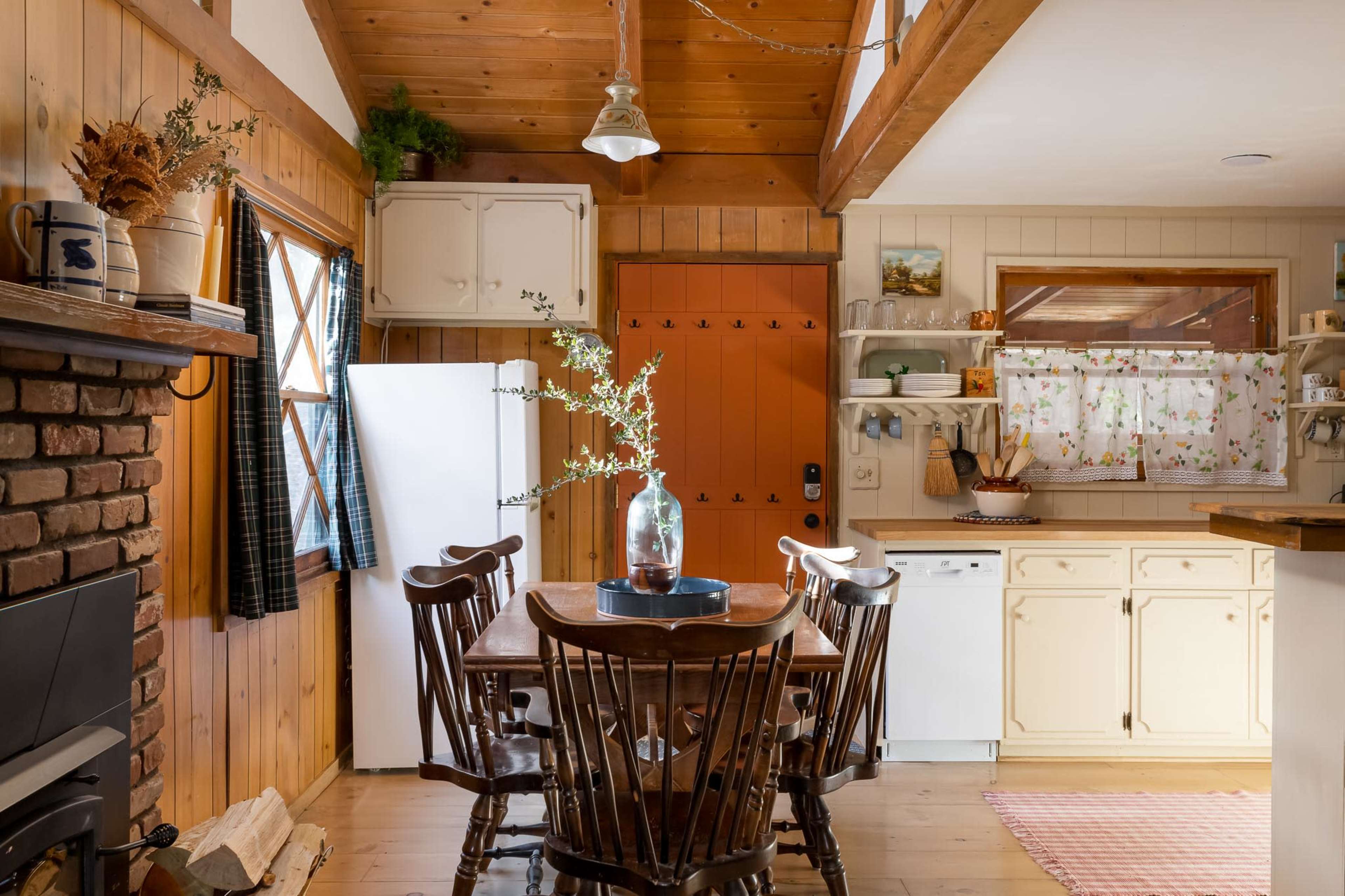A cozy kitchen features a wooden dining table surrounded by chairs, a white refrigerator, and an orange door, with warm wood paneling and a brick fireplace in the background.
