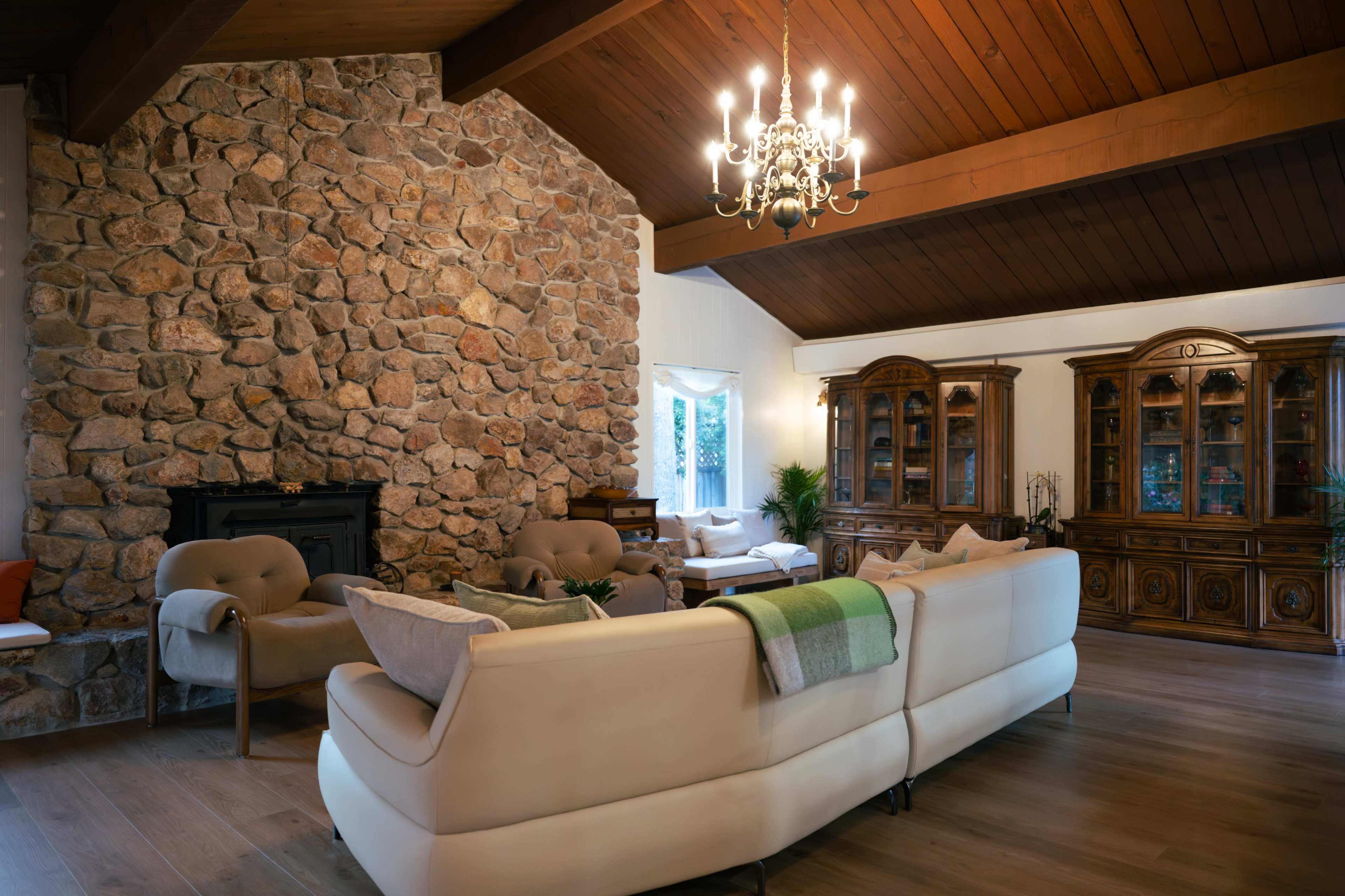 The image shows a spacious living room with a stone fireplace, a high wooden ceiling, and elegant wooden furniture along with a cozy seating area.