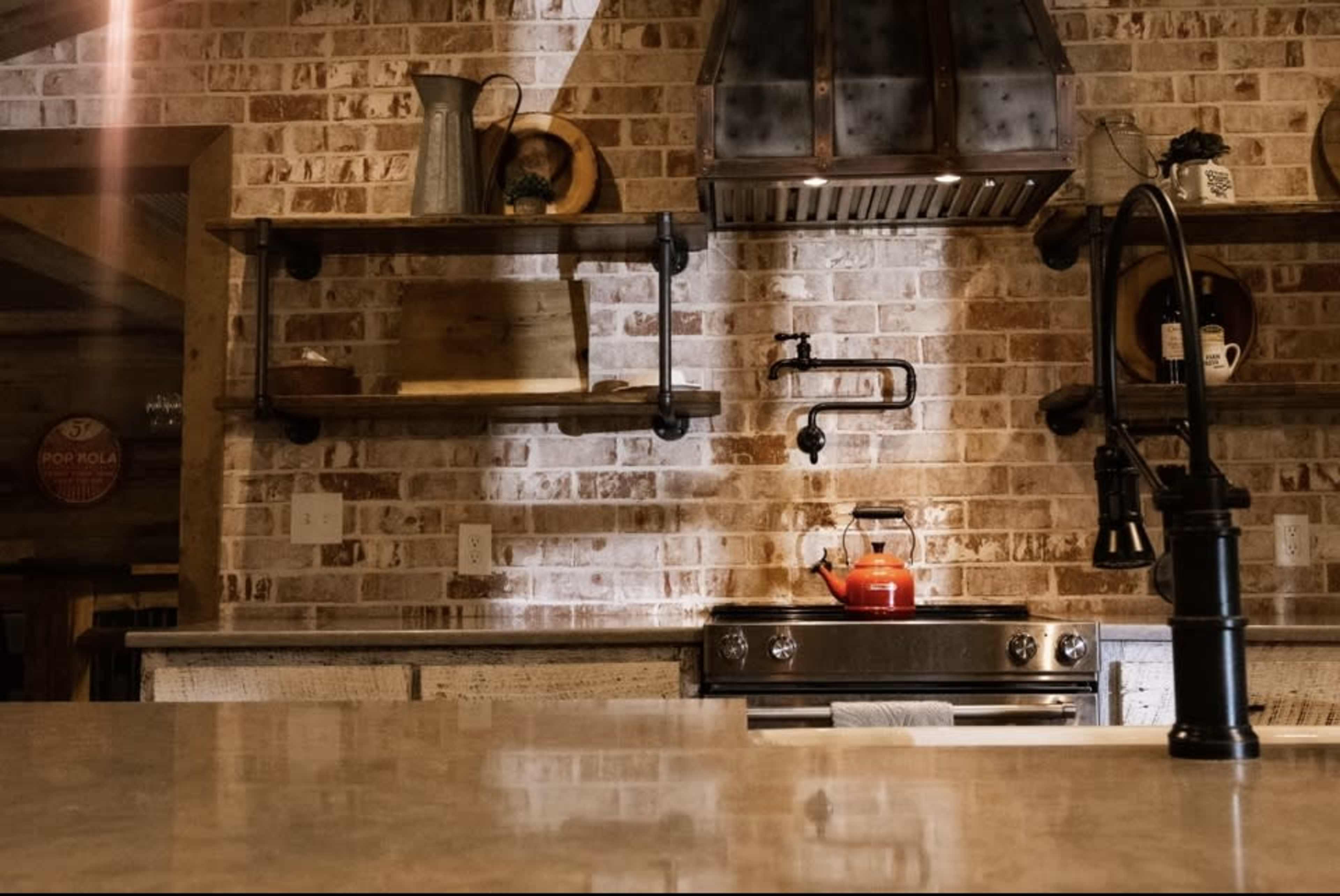A rustic kitchen with exposed brick walls, wooden shelves, and a stainless steel stove beneath a metallic range hood.