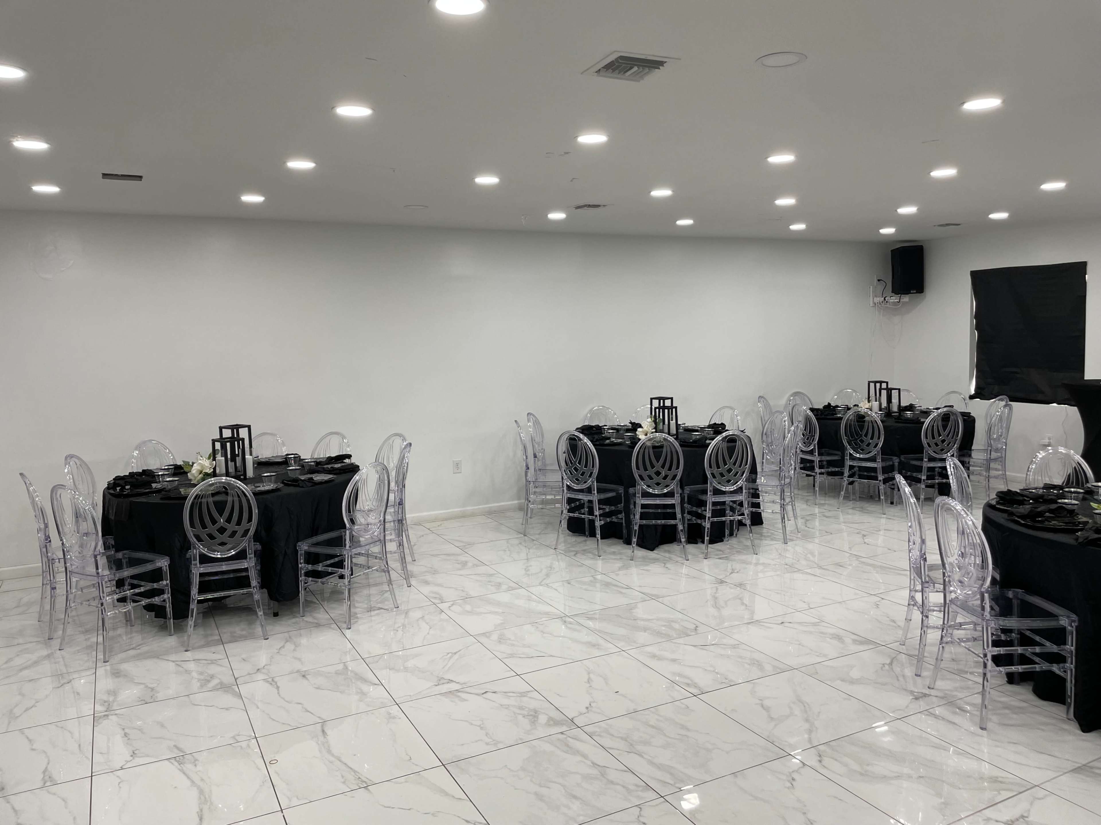 A spacious event room features several tables covered with black tablecloths and surrounded by transparent chairs, with white walls and marble flooring.