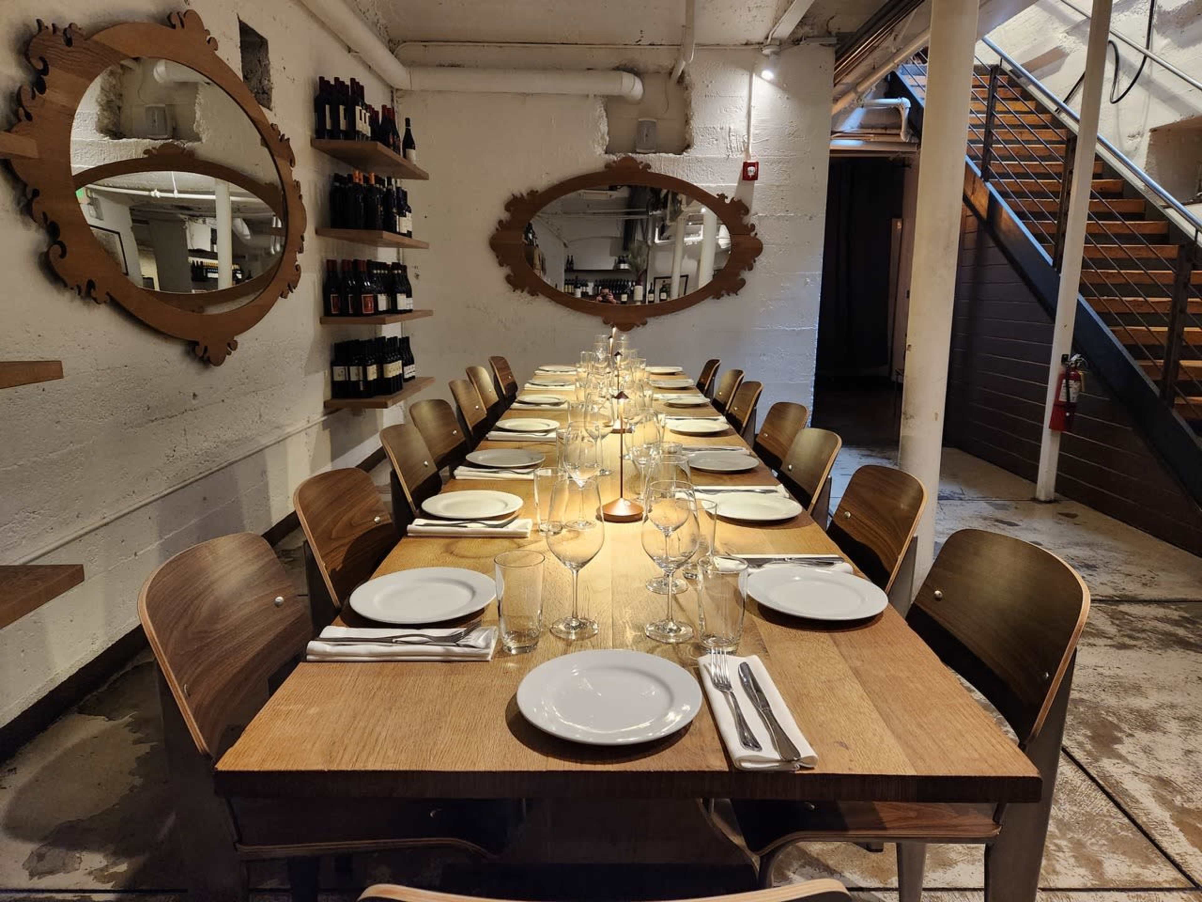 A long dining table is set with plates and glassware in a well-lit restaurant cellar, surrounded by wooden chairs and mirrors on the walls.
