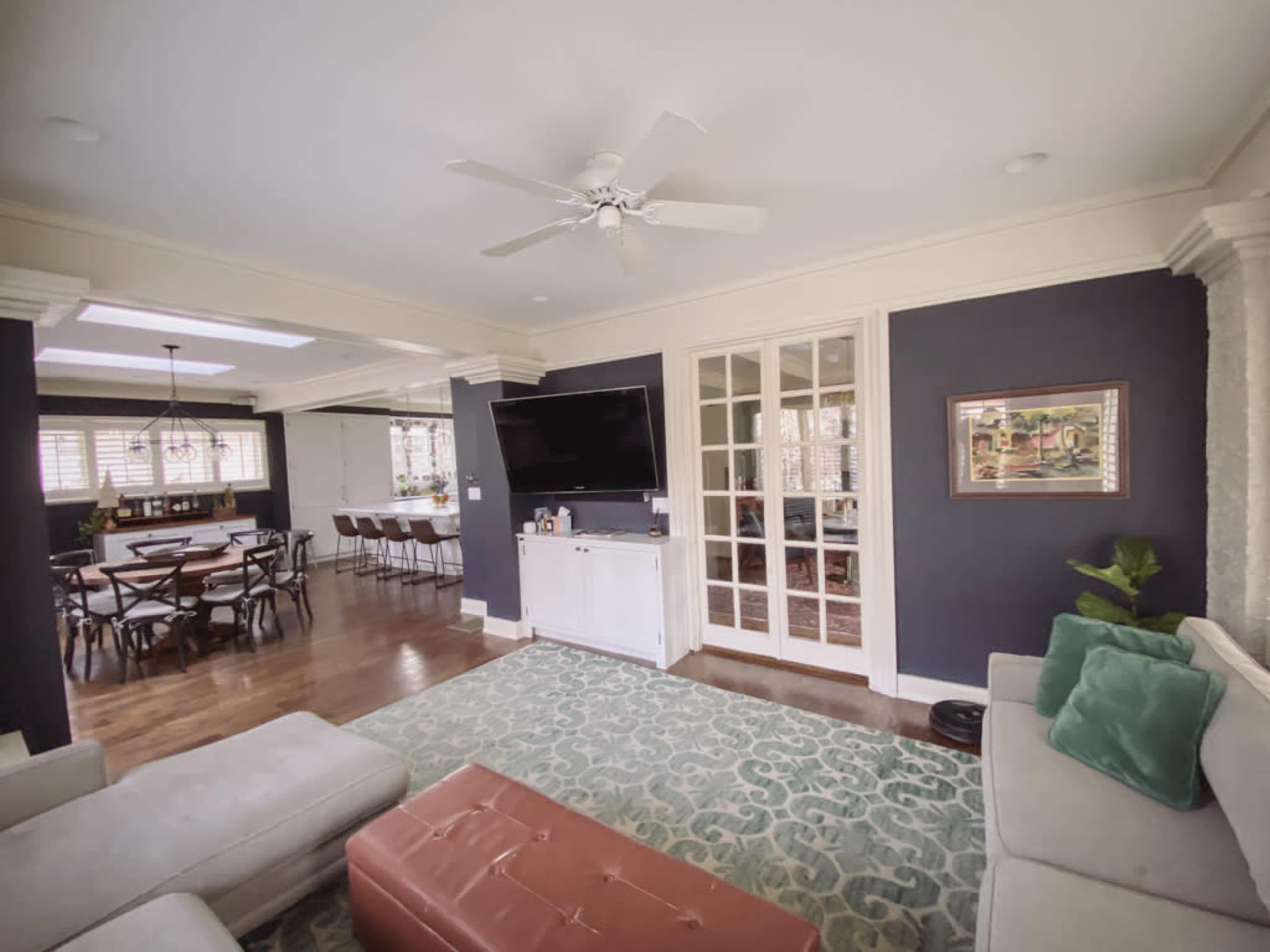 The image shows a living room with a ceiling fan, a television on the wall, and a view into a dining area featuring a table and chairs.