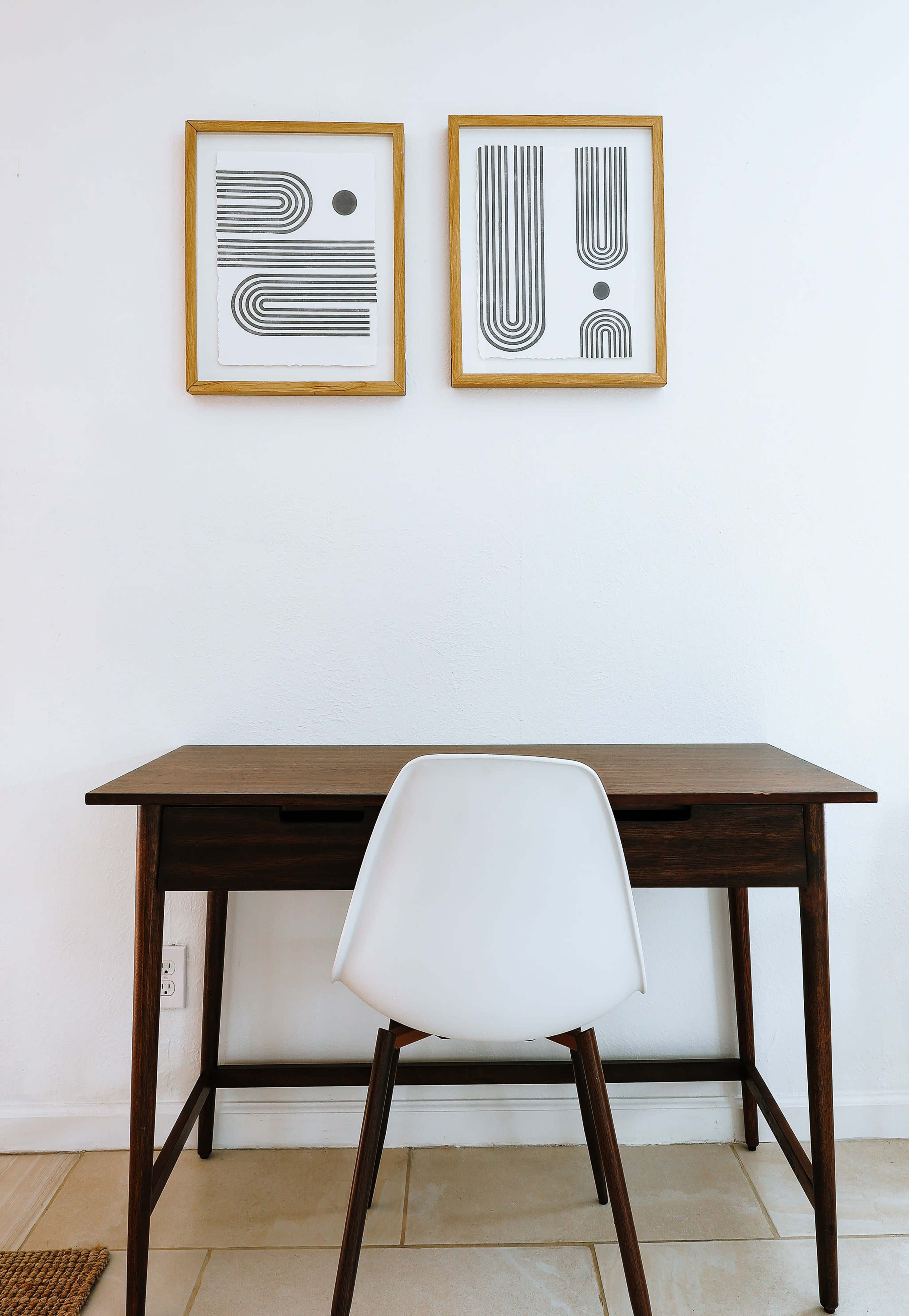 A wooden desk with a white chair is positioned against a white wall, featuring two framed geometric prints above it.