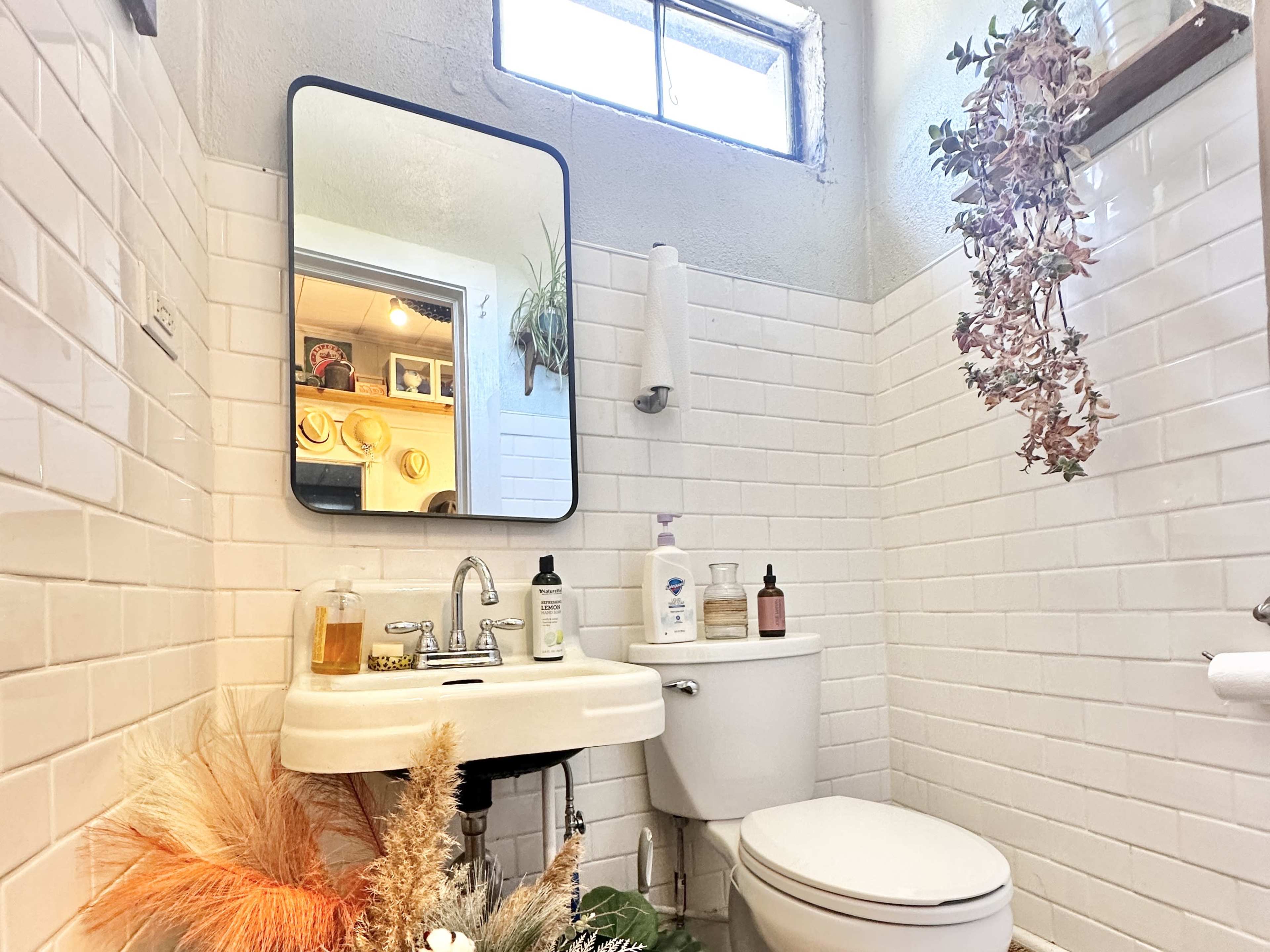 A small bathroom featuring a white tiled wall, a mirror above a sink, and a toilet, with a window allowing natural light and decorative plants on the wall.