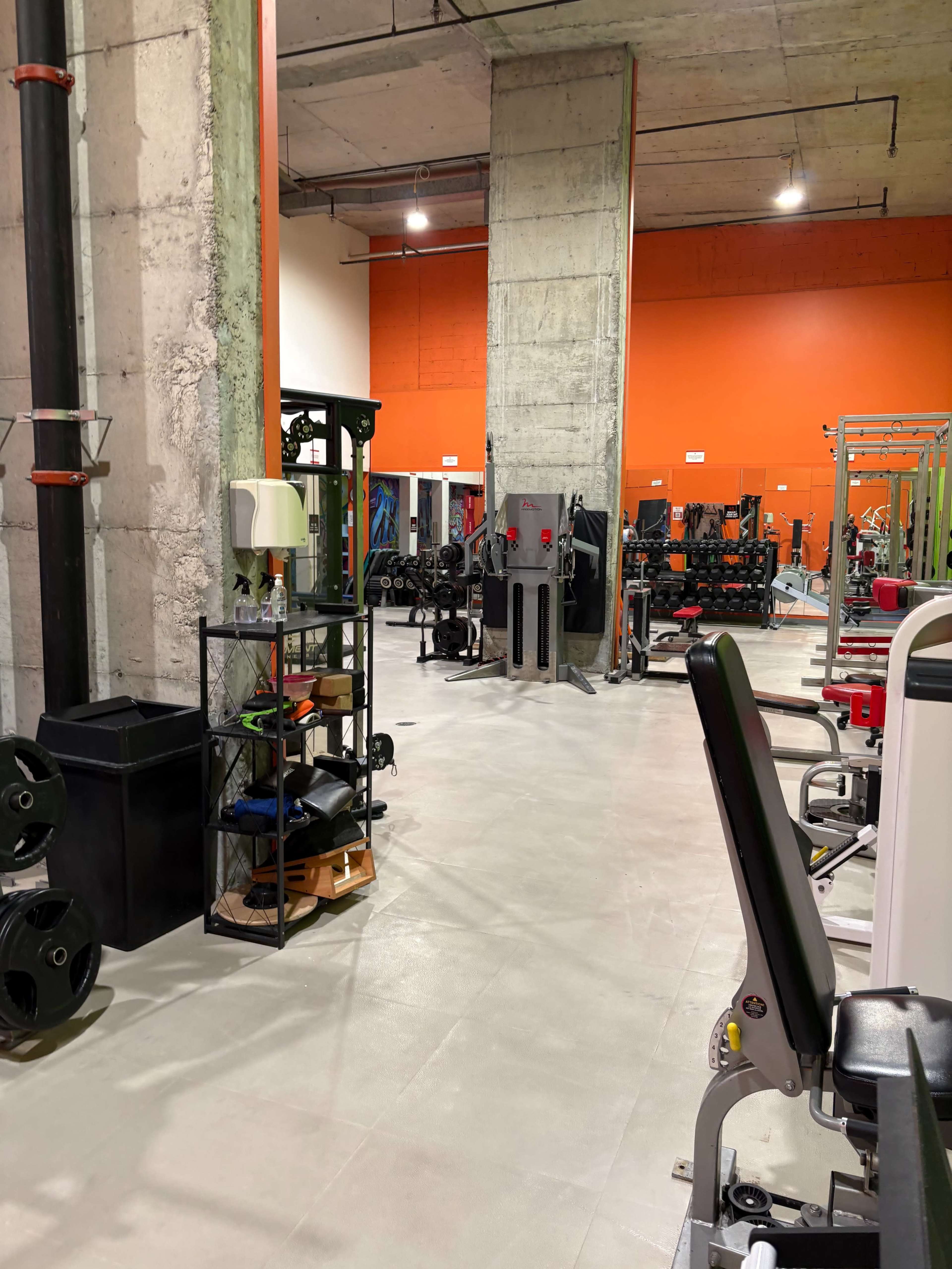 The image shows a spacious gym interior featuring various exercise equipment, including weight racks and machines, set against a brightly colored orange wall.