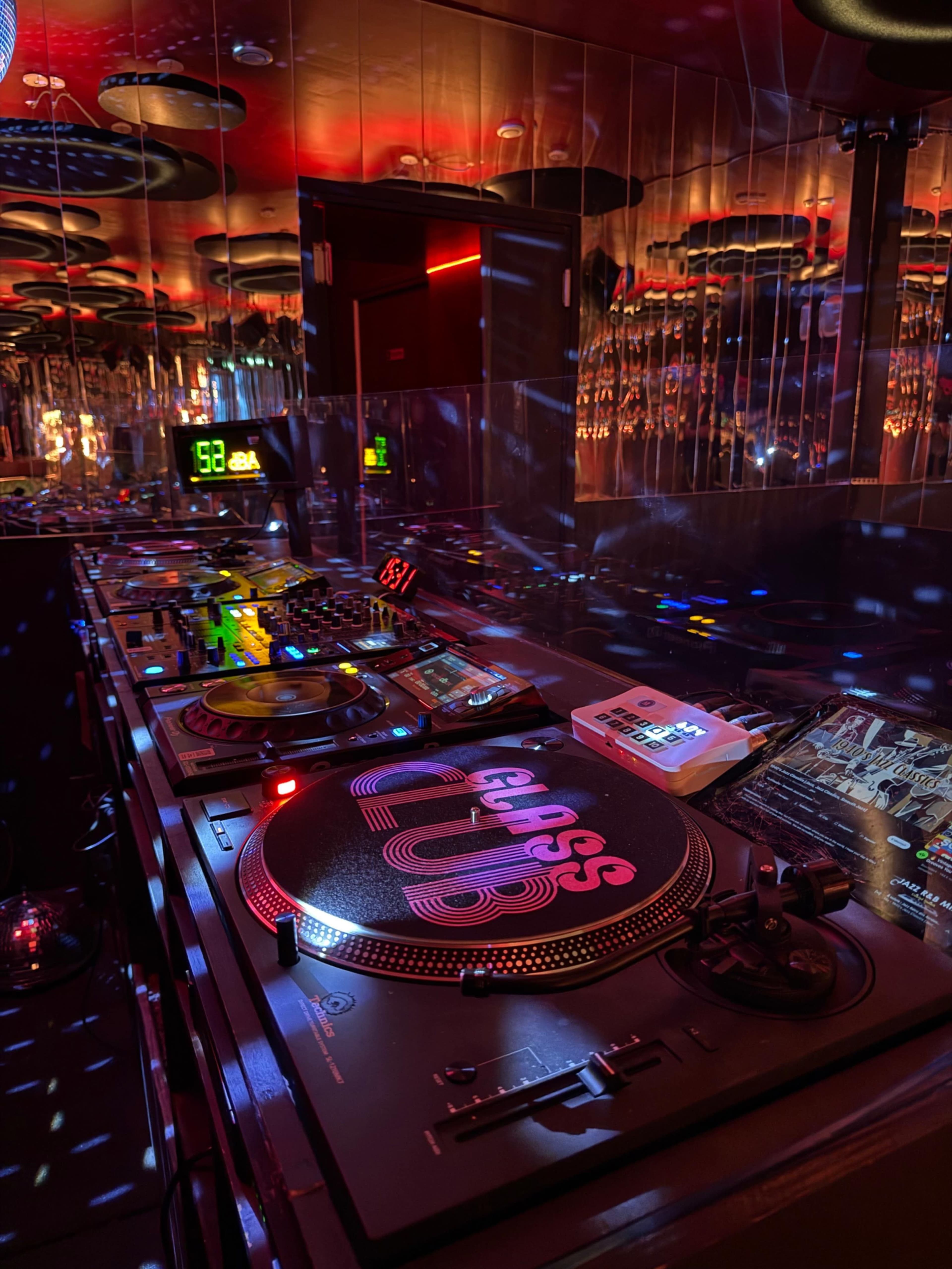 A DJ booth is set up with turntables, mixers, and lighting in a vibrant, mirrored club environment.