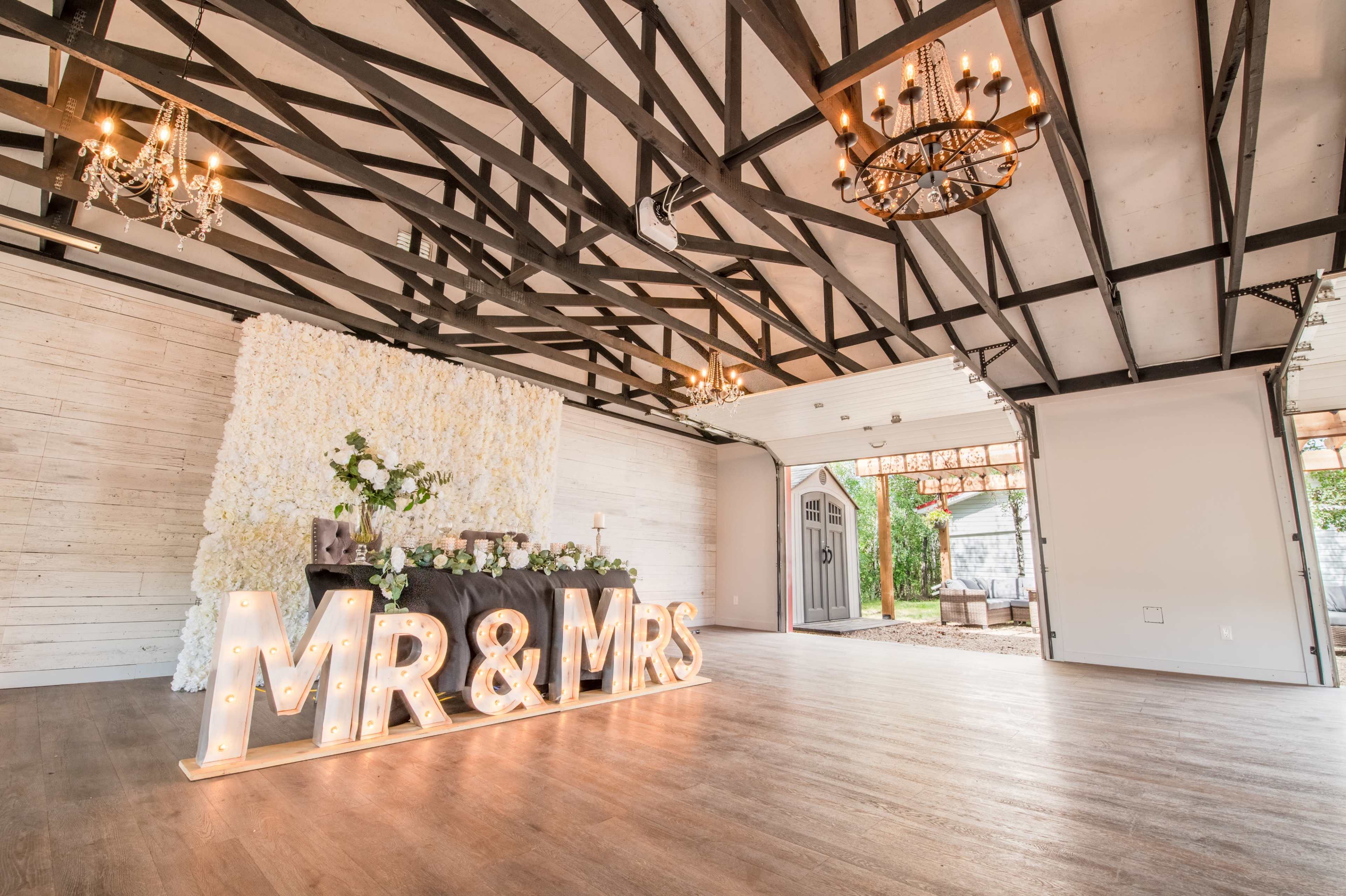 A spacious event venue features a decorative backdrop of flowers, a table set for a celebration, and large illuminated letters spelling "MR & MRS."