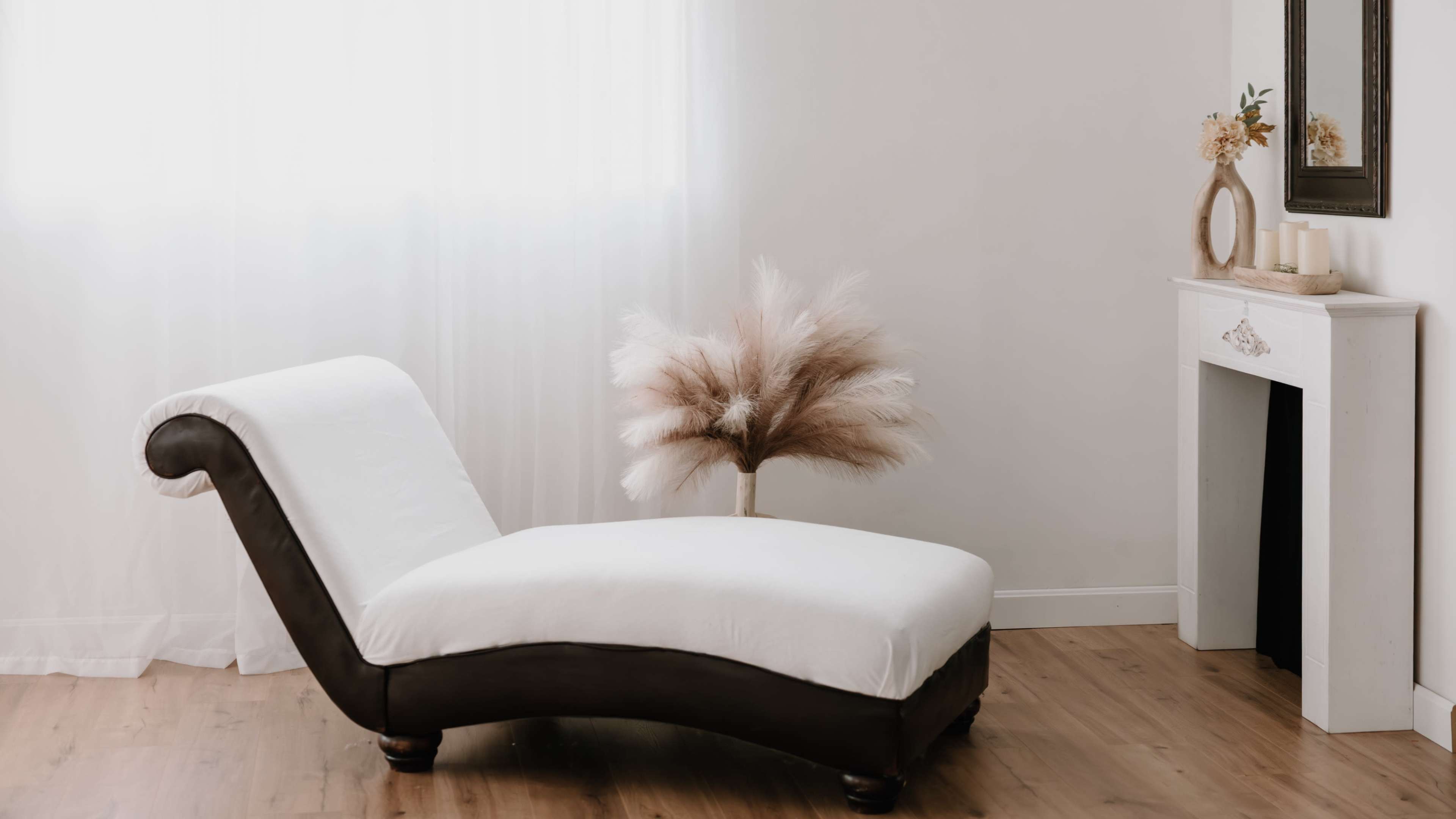 A lounge chair with a white cushion and dark wooden frame sits next to a tall pampas grass arrangement in a bright, minimalist room.