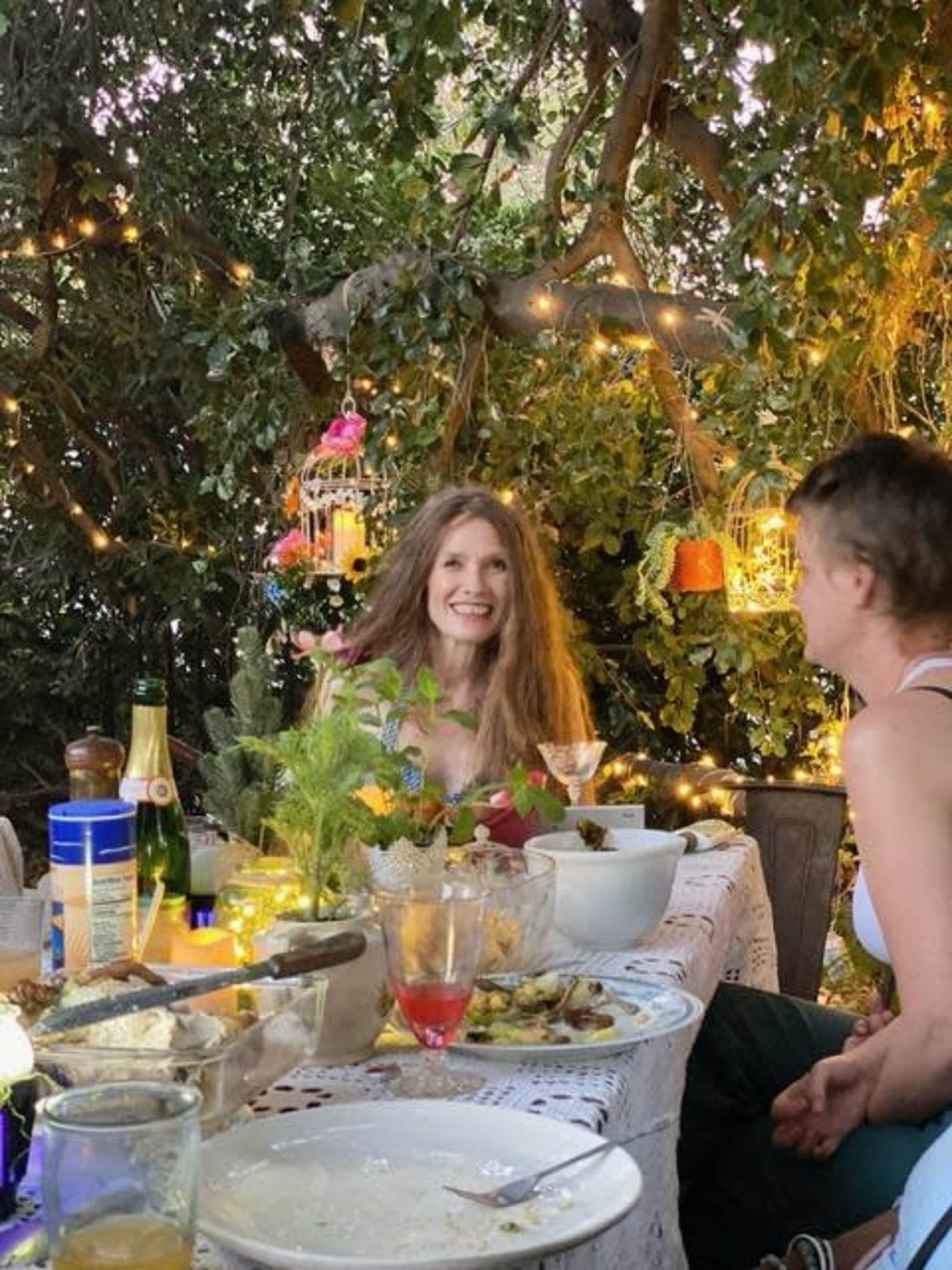 A woman with long hair sits at a table adorned with food and drinks, surrounded by greenery and decorative lights.