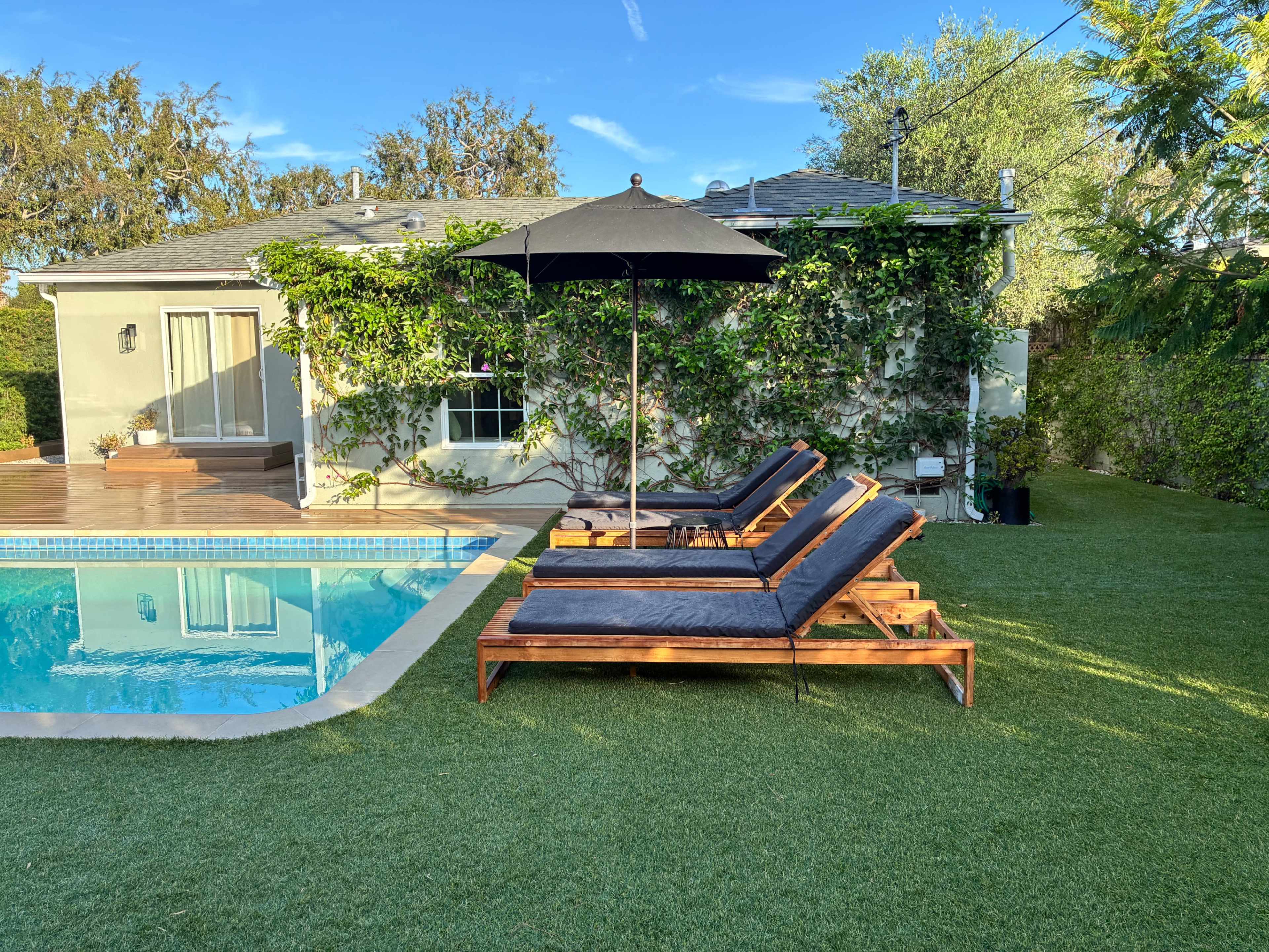 The image shows two lounge chairs with an umbrella beside a swimming pool in a backyard with green grass and a house covered in foliage in the background.