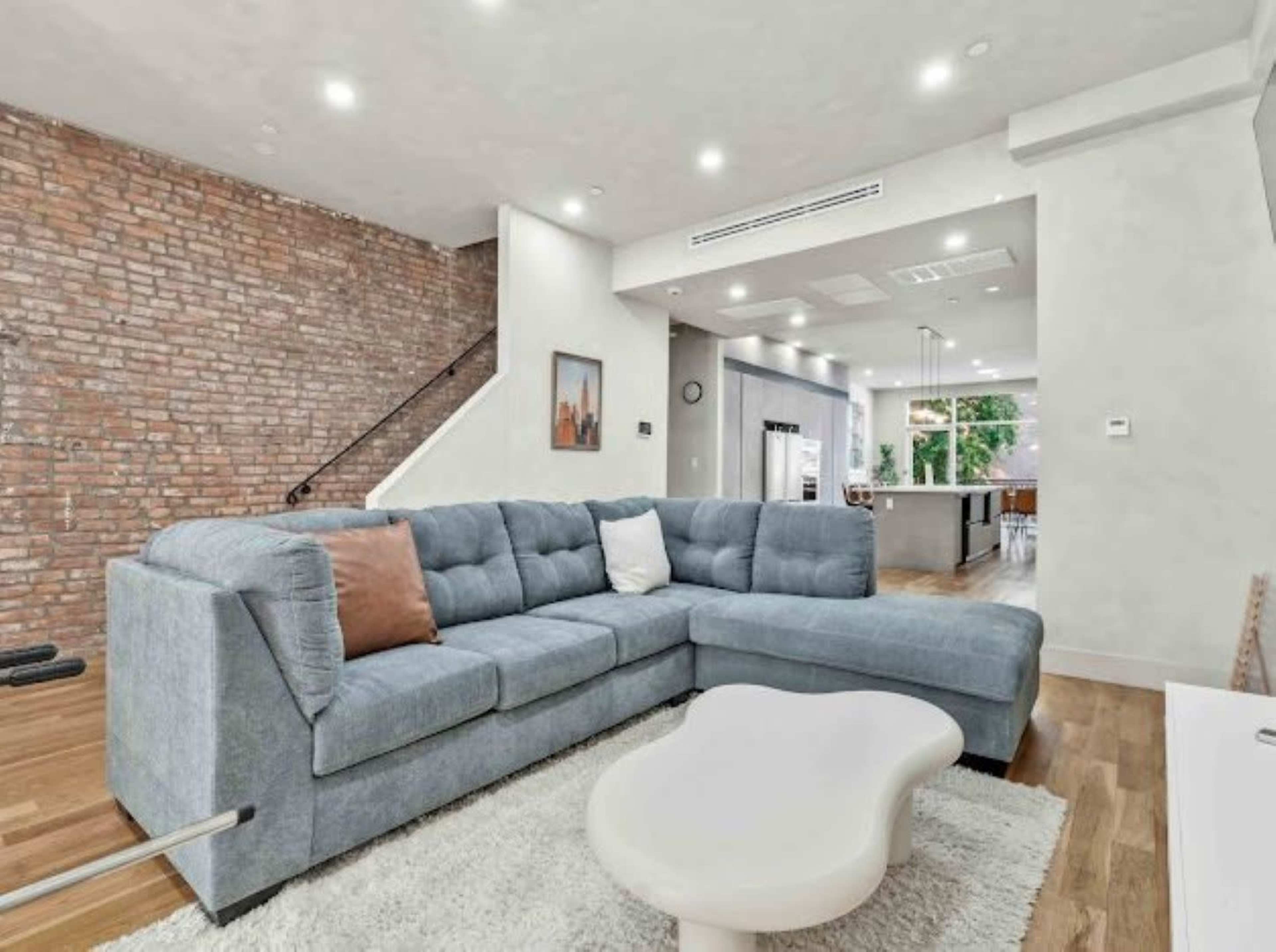 The image shows a modern living room featuring a blue sectional sofa, a light rug, exposed brick walls, and an open floor plan leading to a kitchen area.