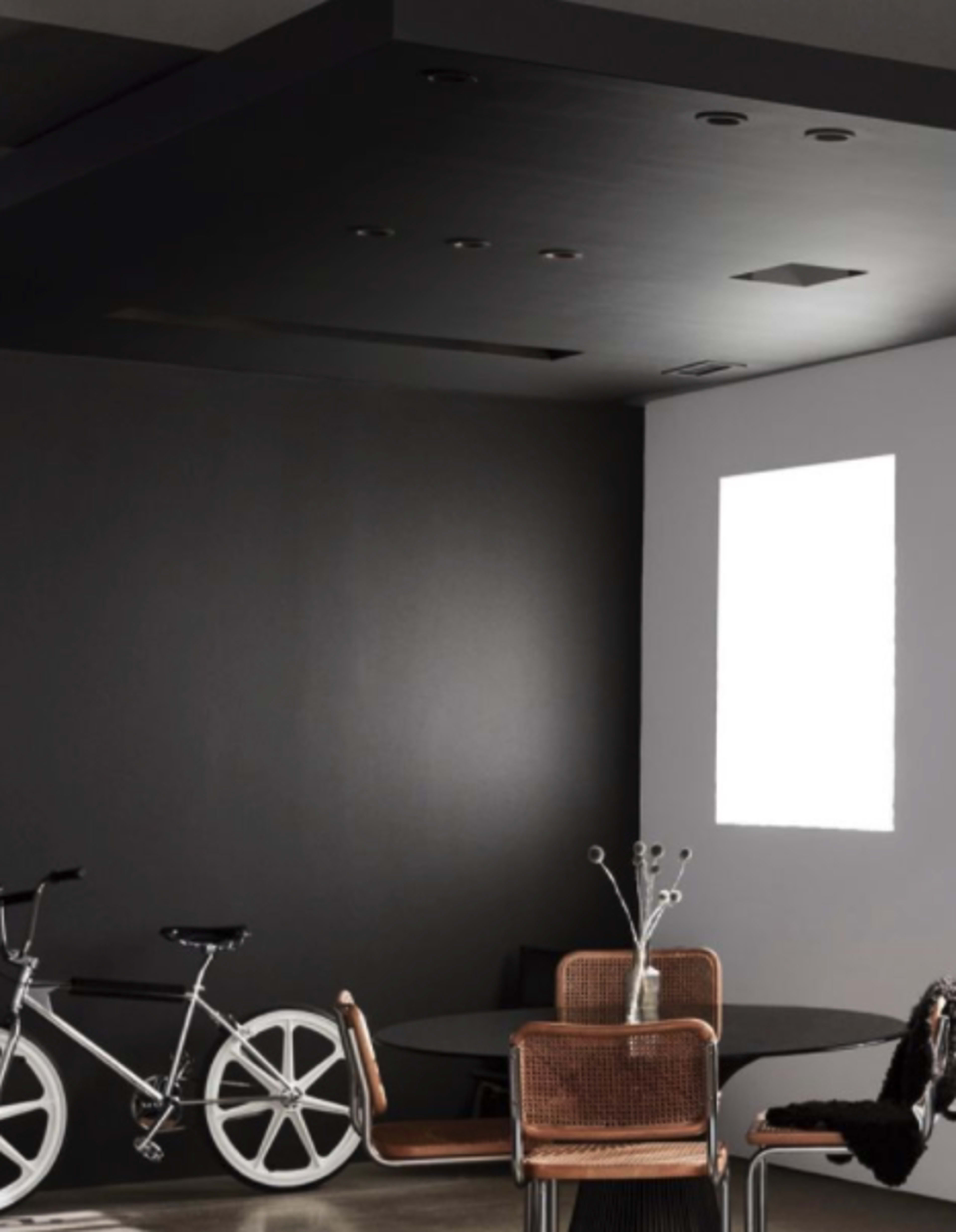 A modern interior features a black wall with a white rectangular window, a round table with woven chairs, and a bicycle leaning against the wall.