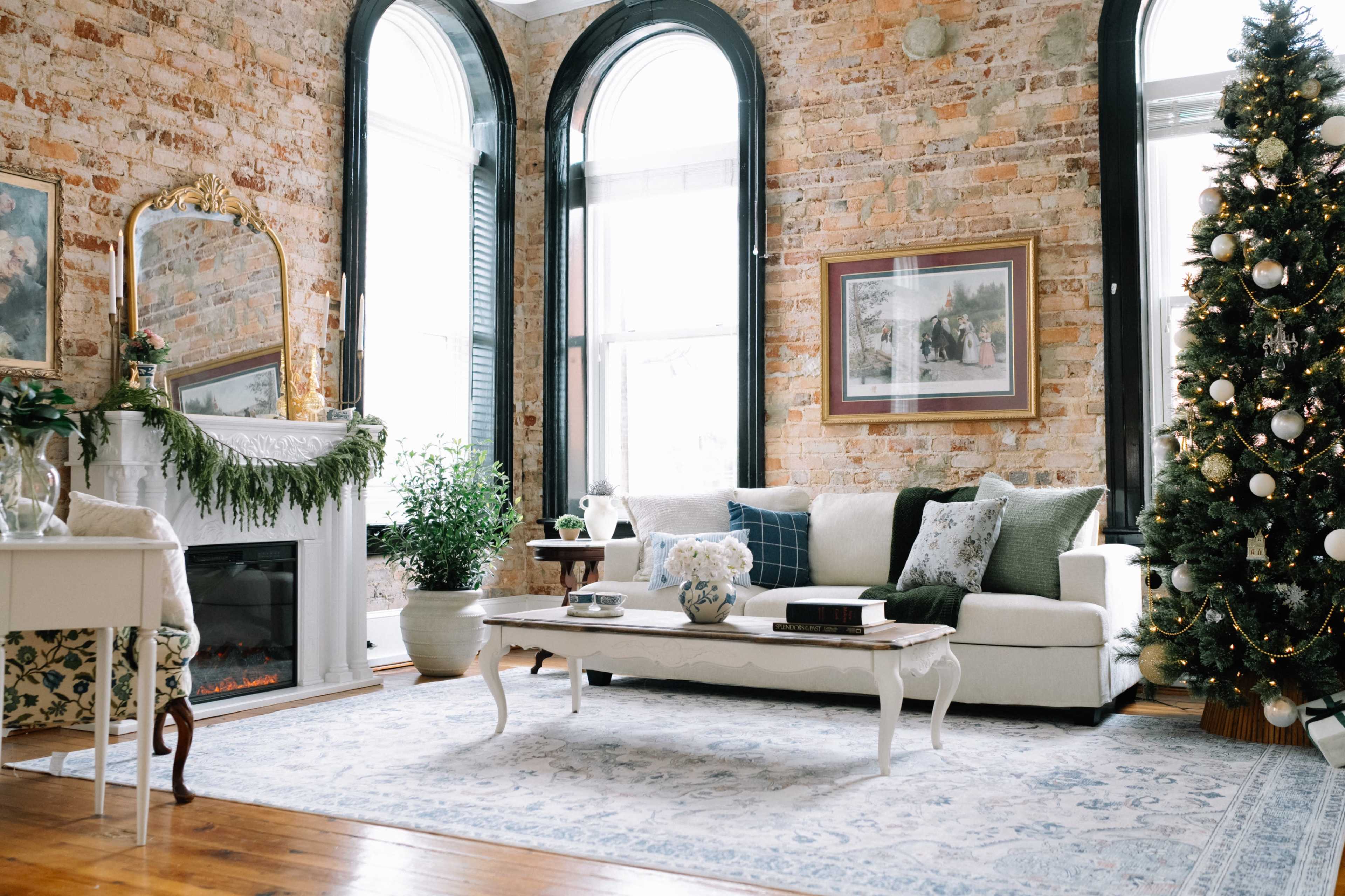 The living room features a white sofa, a coffee table, a decorative fireplace, tall windows, and a Christmas tree adorned with ornaments.